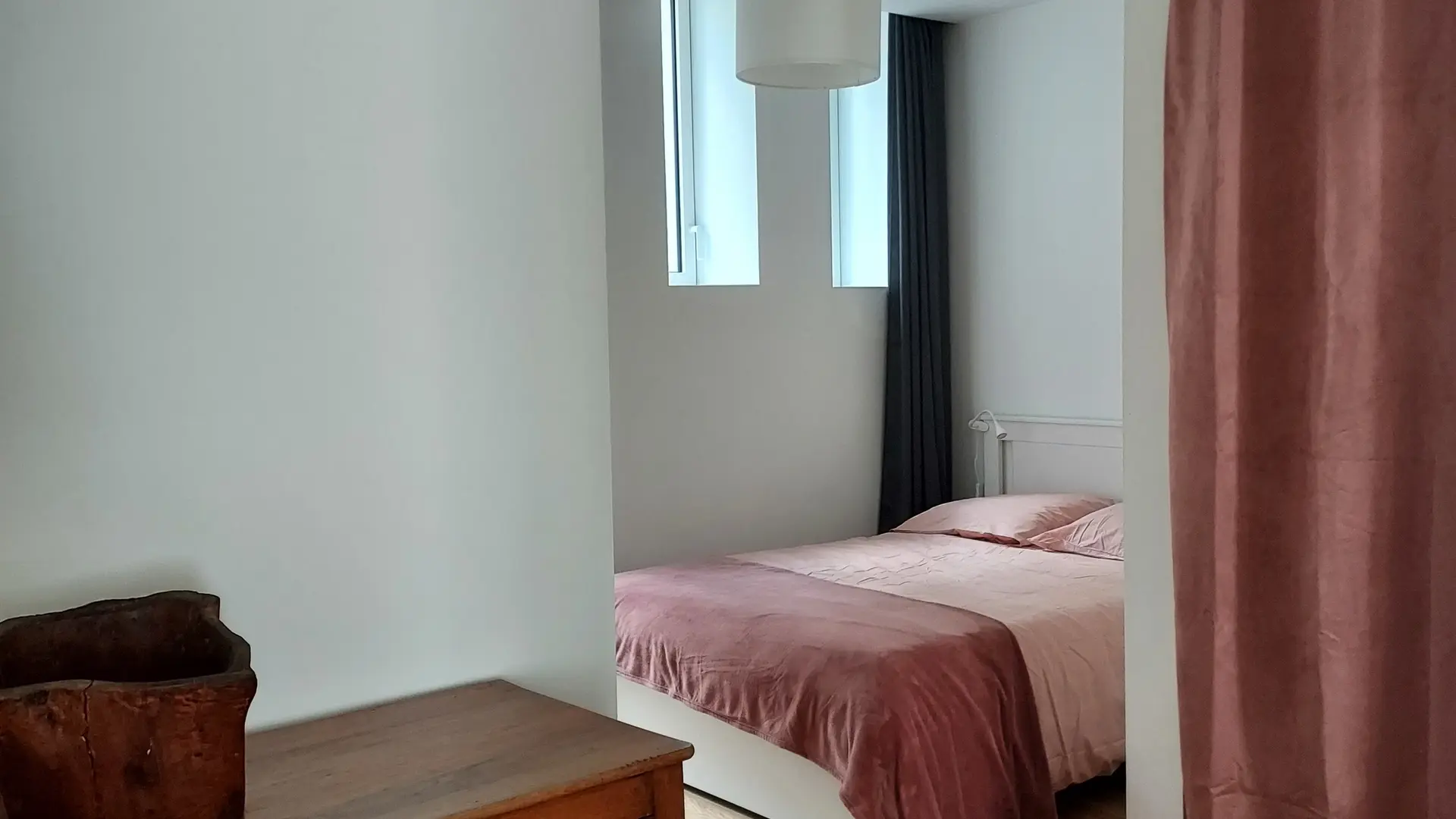 La chambre rose avec un grand lit recouvert d'une couette rose. Un rideau rose sépare la chambre du reste de la pièce. On peut voir un meuble en bois au premier plan.