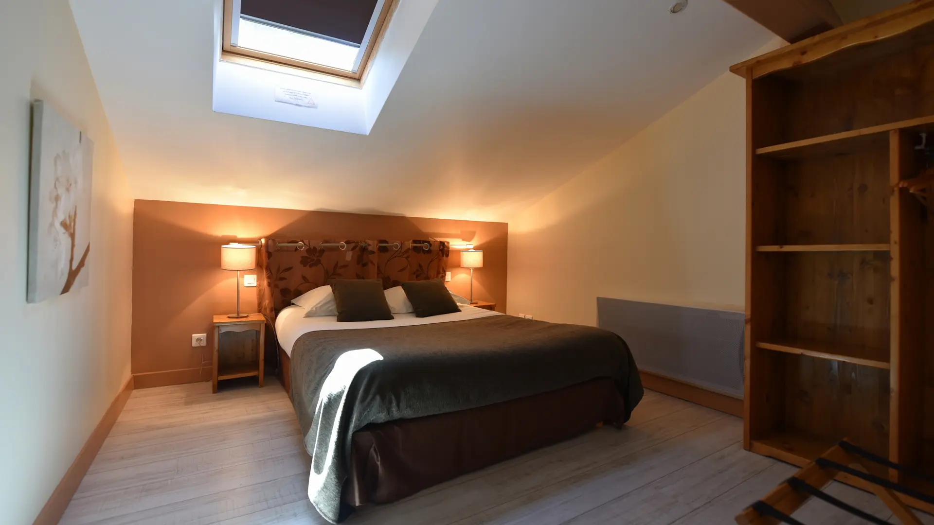 Chambre dotée d’un lit double, de deux tables de chevet avec lampes allumées, d’un vélux laissant entrer la lumière naturelle, d’un cadre mural, d’une armoire ouverte et d’un porte-bagage. Un radiateur est aussi visible dans la pièce