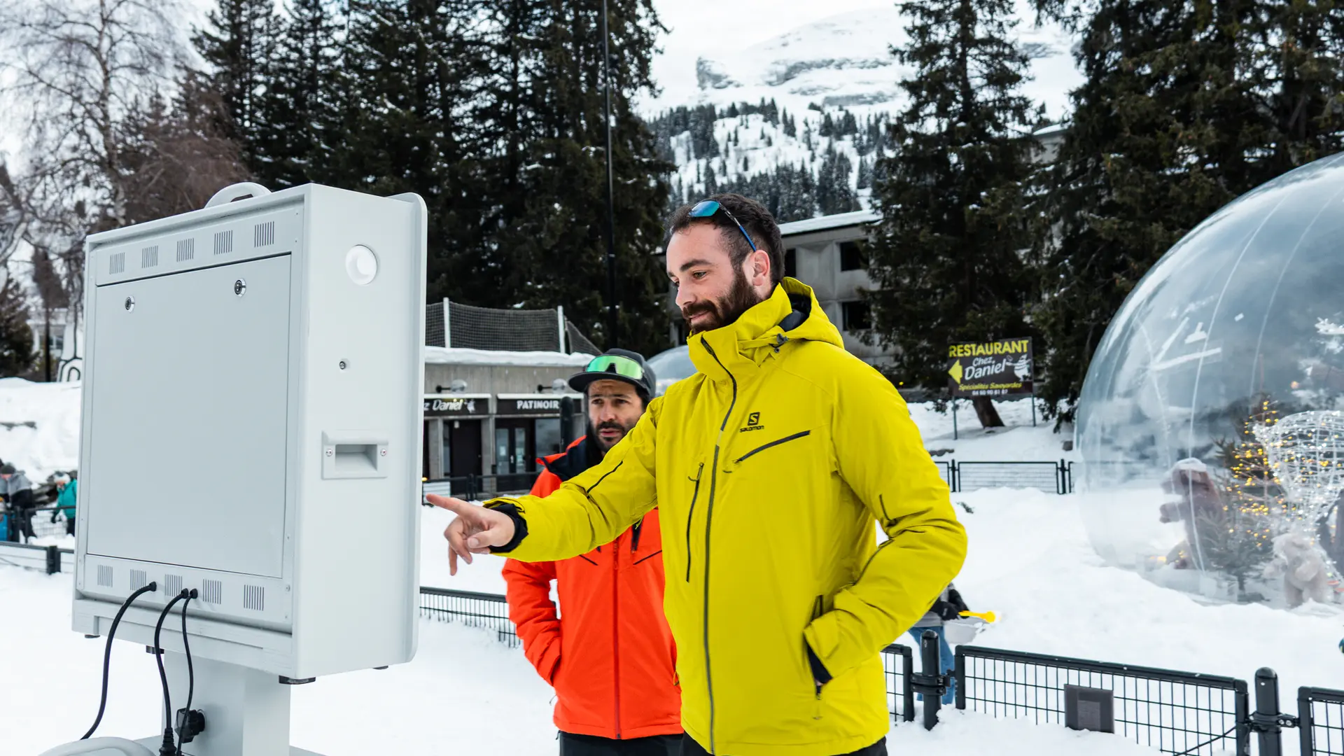 Deux hommes en tenue de ski utilisent une borne photo interactive dans une station enneigée, l'un pointant l'écran du doigt. Ils se trouvent en extérieur, entourés de sapins, d'une bulle décorative géante et de bâtiments de montagne.