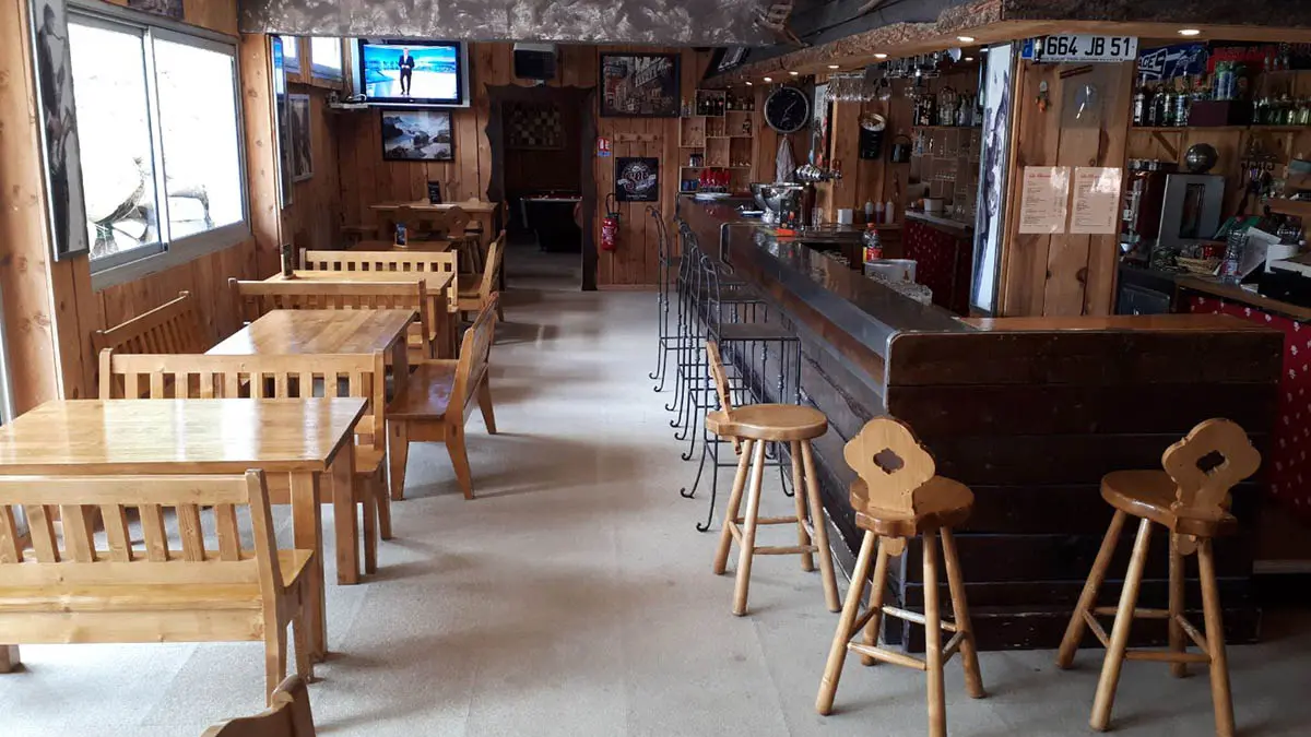 Salle d'accueil du restaurant, comptoir bar en bois laqué, tables et chaises, tabourets, salle de jeux au fond, TV, fenêtres donnant sur l'extérieur