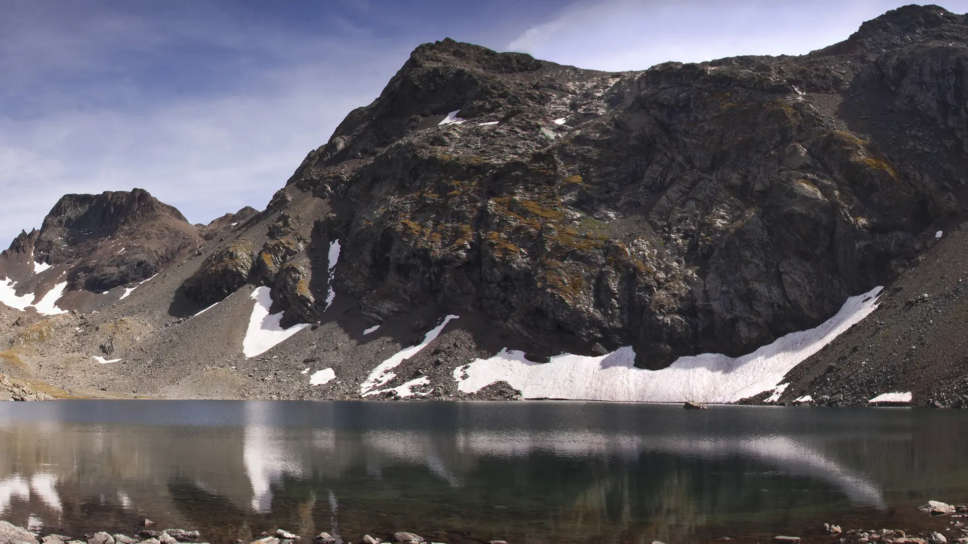 Le paysage est composé d'un lac tranquille entouré de montagnes rocheuses et abruptes, avec des traces de neige sur les sommets.