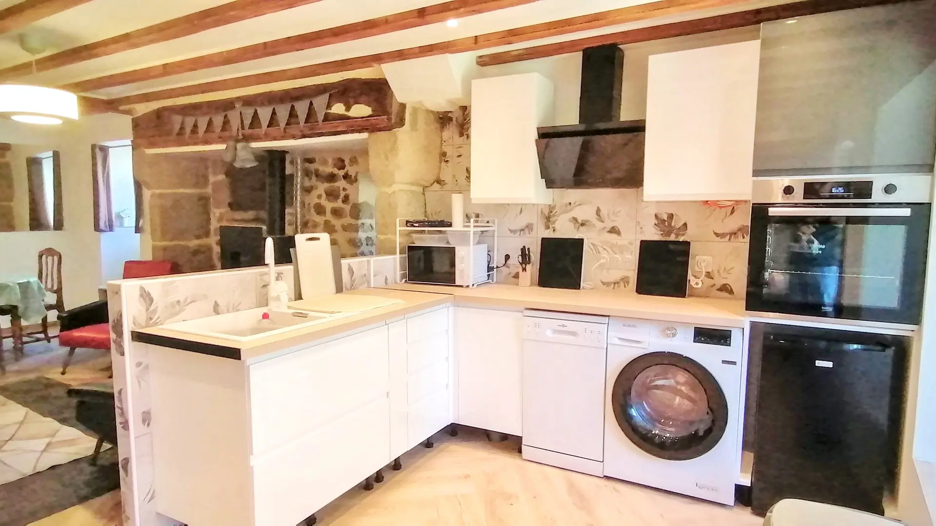 Une cuisine moderne en forme de L dans un espace ouvert. Les armoires et les tiroirs inférieurs sont blancs et le plan de travail est de couleur bois clair. La cuisine comprend un évier encastré, un lave-vaisselle et un lave-linge séchant encastré en façade. Le mur derrière le plan de travail est recouvert d'un carrelage décoratif clair avec des motifs discrets. Les appareils électroménagers sont noirs et gris, et comprennent un four encastré, une hotte aspirante au design moderne, et un petit réfrigérateur noir sous le four. Un four à micro-ondes blanc est posé sur le plan de travail. L'espace dispose de poutres en bois apparentes au plafond. À l'arrière-plan, une arche révèle un mur en pierres apparentes rustiques. Au-delà, on aperçoit un coin repas avec une table et des chaises en bois, ainsi qu'un miroir sur le mur, ce qui indique que la cuisine est ouverte sur le salon ou la salle à manger.