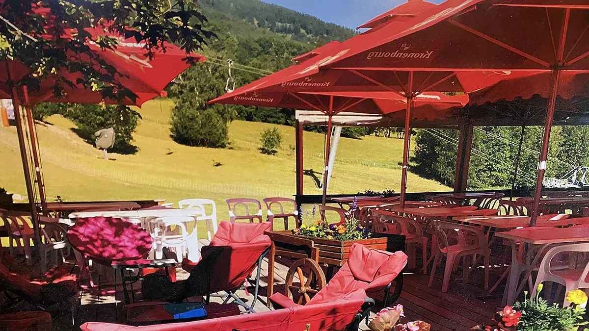 Terrasse extérieure du restaurant en été, tables et chaises en plastique, grands parasols, vue sur un champ et sur les montagnes