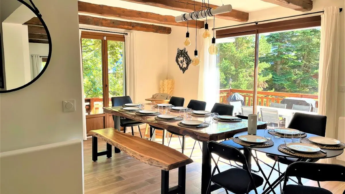 Séjour lumineux avec table à manger, ameublement bois, poutres au plafond, portes-fenêtres donnant sur une terrasse en bois