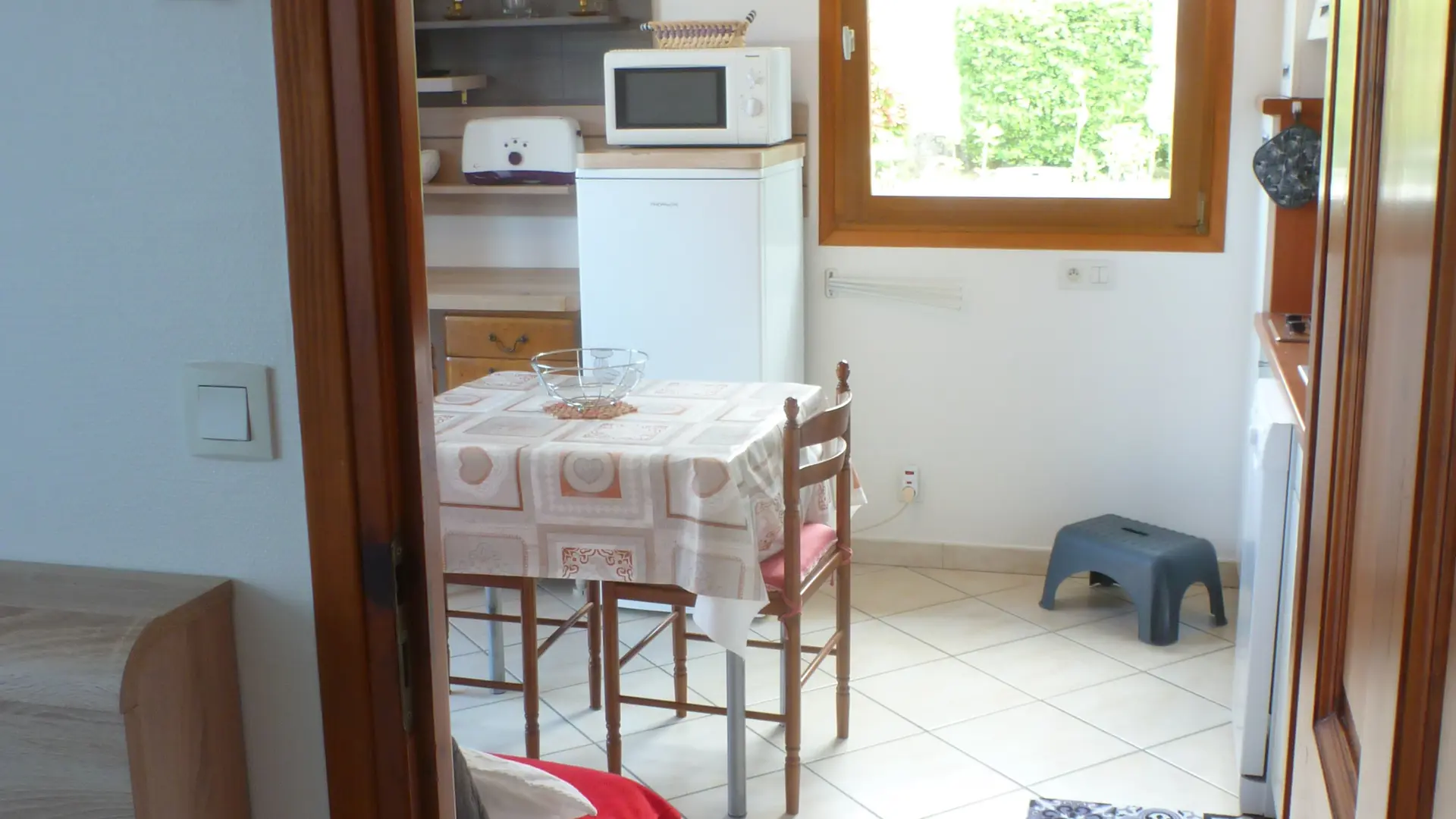Depuis la chambre, on aperçoit la pièce à vivre avec une petite cuisine et un coin repas visible contenant une table recouverte d'une nappe, deux chaises, un petit réfrigérateur, un micro-ondes, et une fenêtre qui donne sur le jardin.
