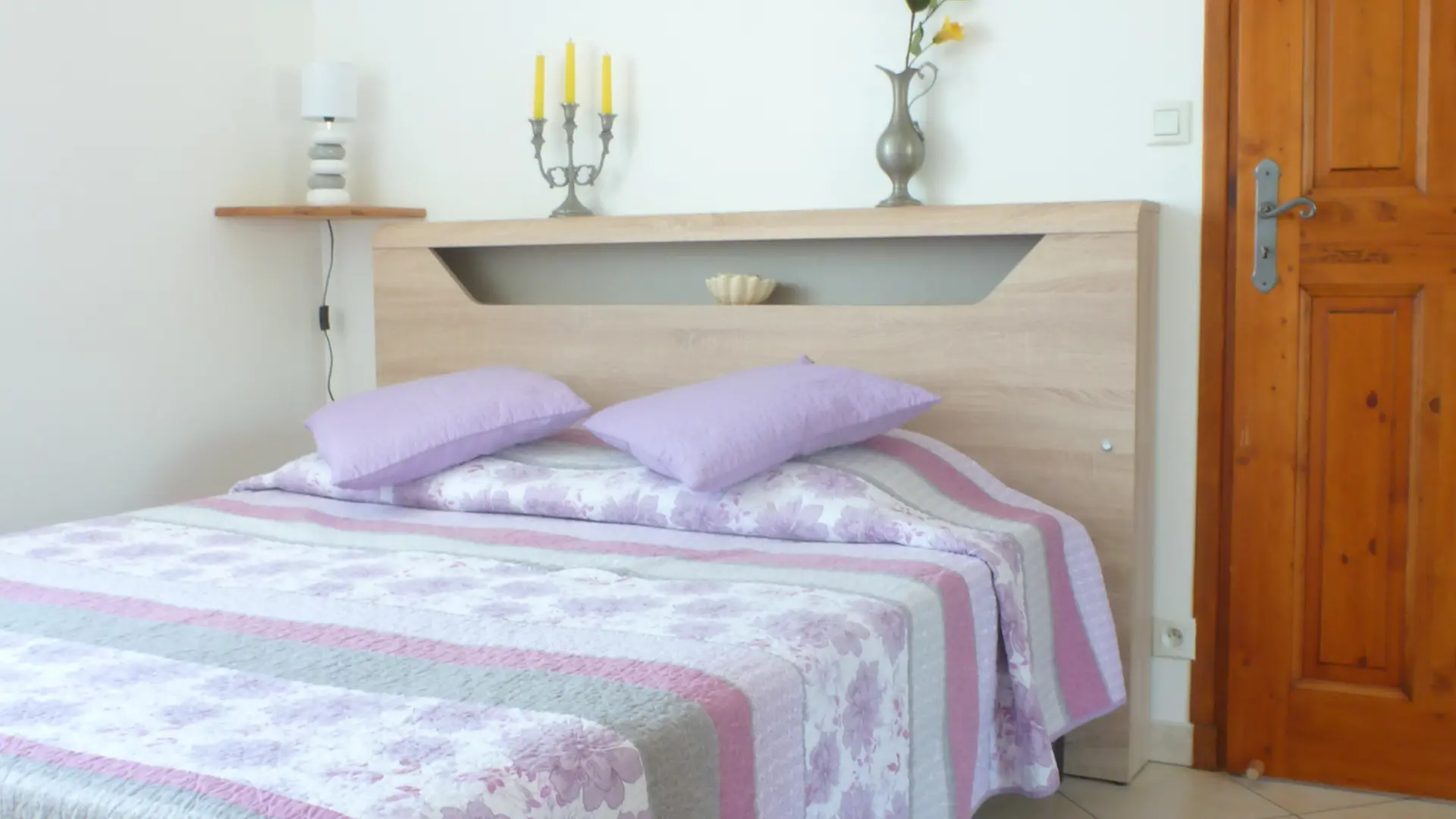 La pièce est une chambre sobre et lumineuse contenant un grand lit avec une parure violette et blanche, une tête de lit claire surmontée d'un tableau encadré, et une porte en bois fermée sur la droite.