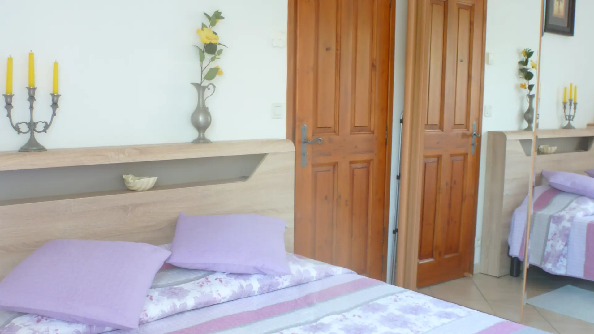 La pièce est une chambre double avec des murs blancs, un lit recouvert d'une parure à motifs violets et blancs, une tête de lit décorée de bougeoirs et de vases, et un placard mural avec des portes miroirs.