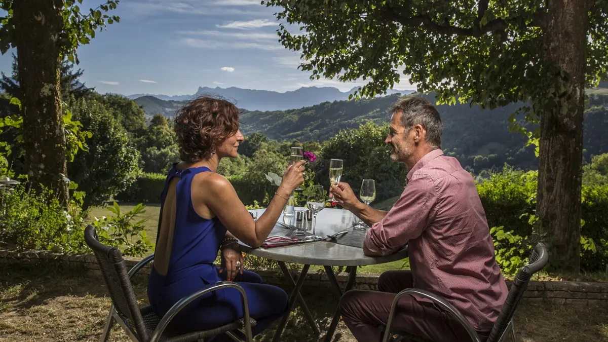 Couple prenant le petit déjeuner sur la terrasse de l'hôtel, sur une table en bois. Derrière eux, un paysage vallonné s'étend à perte de vue. La végétation est luxuriante, avec des arbres aux feuilles abondantes qui créent une atmosphère paisible et naturelle. Le ciel est bleu avec quelques nuages légers.