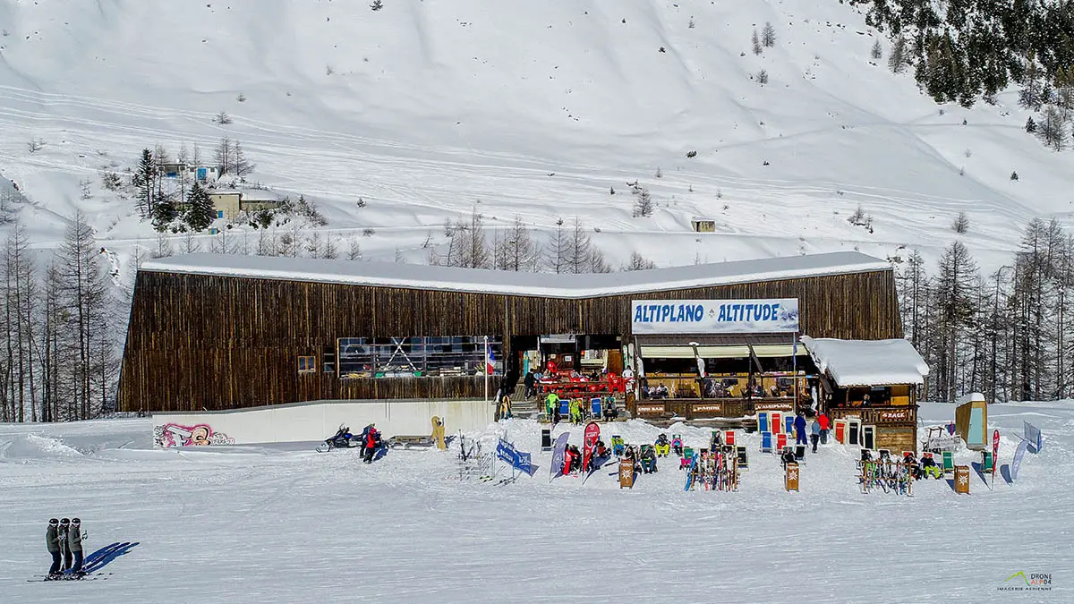 Vue de face du restaurant d'altitude, bâtiment en bois, terrasse extérieure avec chaises longues le long des pistes enneigées, panneau Altiplano - Altitude. Montagnes en arrière-plan