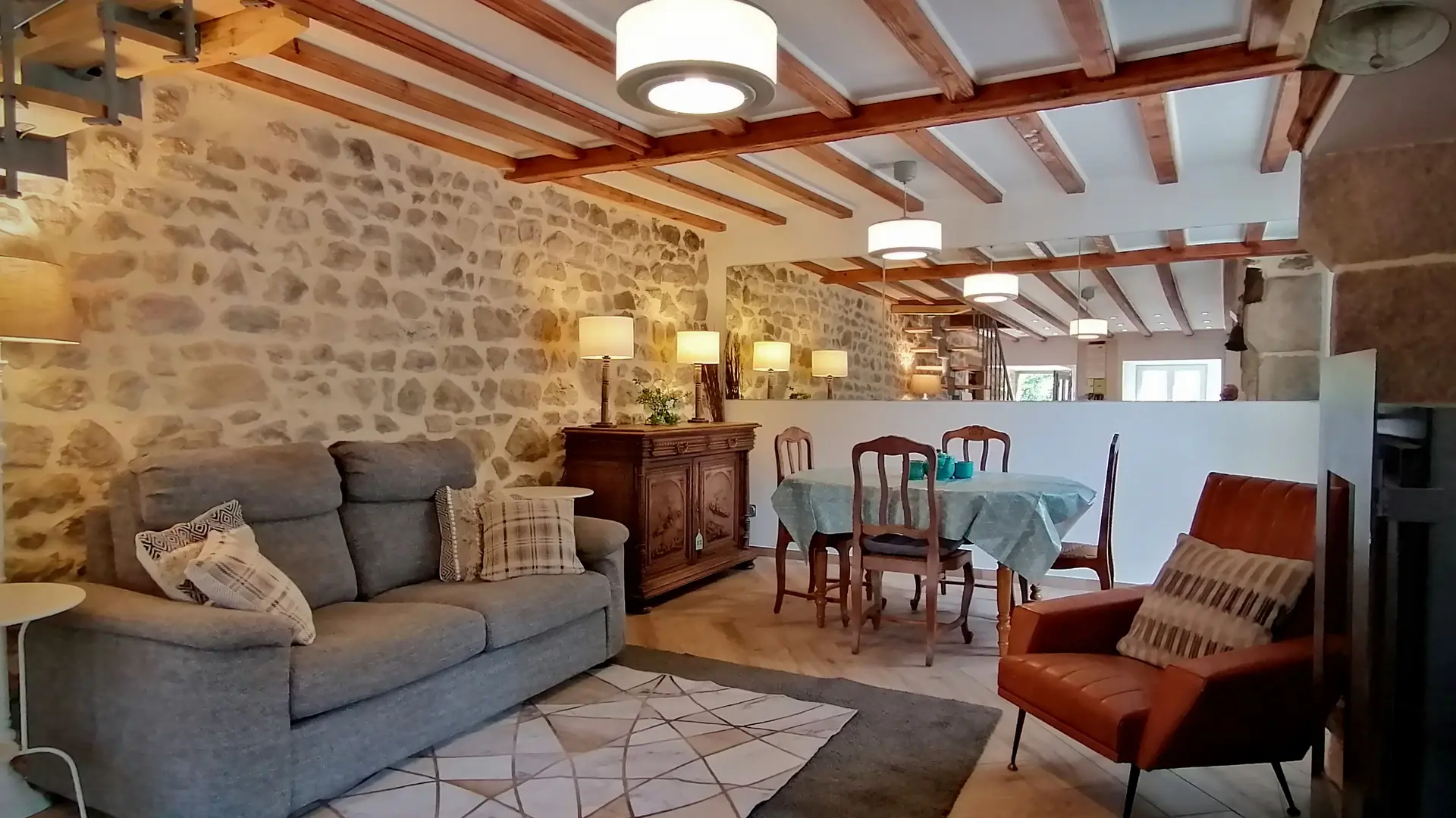 Une pièce de vie comprenant un coin salle à manger et un salon autour d'un poele à bois entouré d'un fauteuil et en face un canapé. La salle à manger comporte un buffet. Divers luminaires offrent un éclairage direct et indirect. Un miroir sur le mur du fond reflète la lumière de la pièce. On voit à droite l'escalier accédant aux chambres.