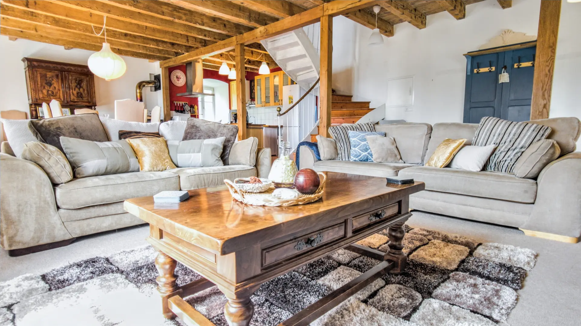 Grand salon chaleureux avec poutres en bois au plafond, comprenant deux grands canapés beige remplis de coussins aux tons doux. Au centre, une large table basse en bois massif repose sur un tapis texturé. À l’arrière-plan, on aperçoit une cuisine ouverte avec meubles en bois clair, un escalier montant à l’étage, ainsi qu’une armoire ancienne bleu foncé. L’ensemble crée une ambiance confortable, lumineuse et accueillante.