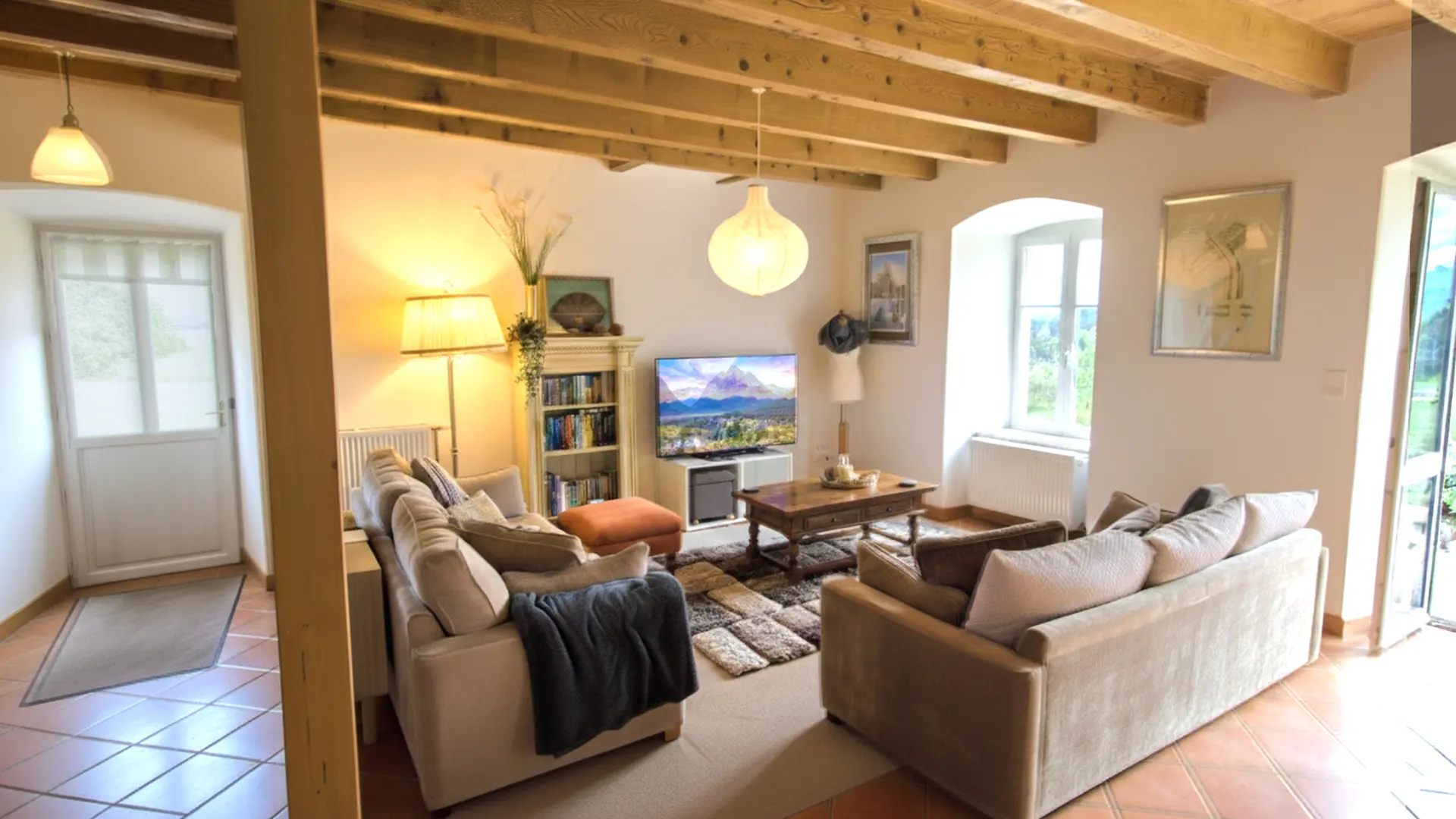 Salon lumineux avec poutres apparentes au plafond, deux grands canapés beige placés face à face autour d’une table basse en bois. Un tapis doux et texturé couvre le sol au centre. Au fond, une bibliothèque garnie de livres se tient à côté d’un téléviseur allumé affichant un paysage de montagne. Une lampe sur pied éclaire l’espace et de grandes fenêtres laissent entrer la lumière naturelle, offrant une vue sur le jardin extérieur. L’ensemble crée une atmosphère chaleureuse, paisible et accueillante.