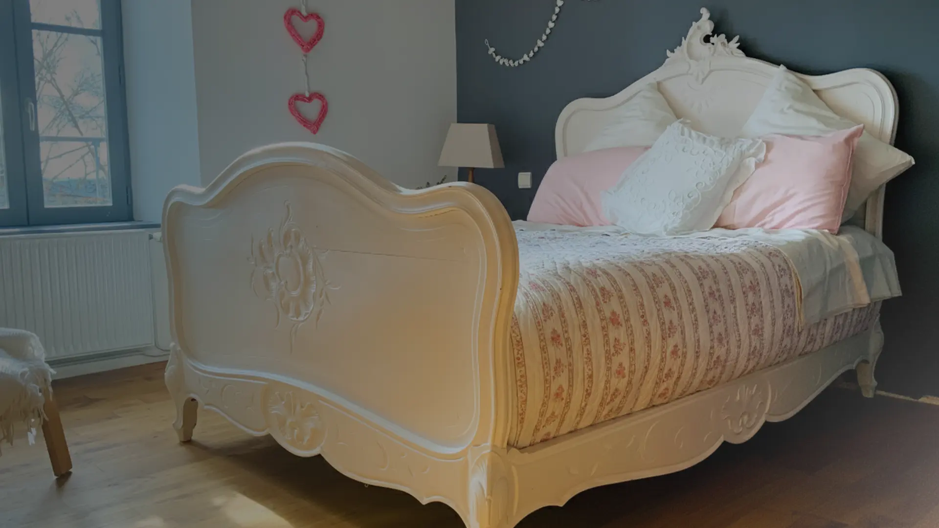 Chambre lumineuse avec un grand lit ancien en bois sculpté de couleur crème. La literie est composée de coussins blancs et roses ainsi que d’une couverture à motifs floraux. Le mur derrière le lit est peint en gris foncé, décoré d’une guirlande et de deux cœurs suspendus. Une lampe de chevet se trouve sur une petite table à côté du lit, et une fenêtre à gauche laisse entrer la lumière du jour. Le sol est en parquet, créant une ambiance douce et romantique.
