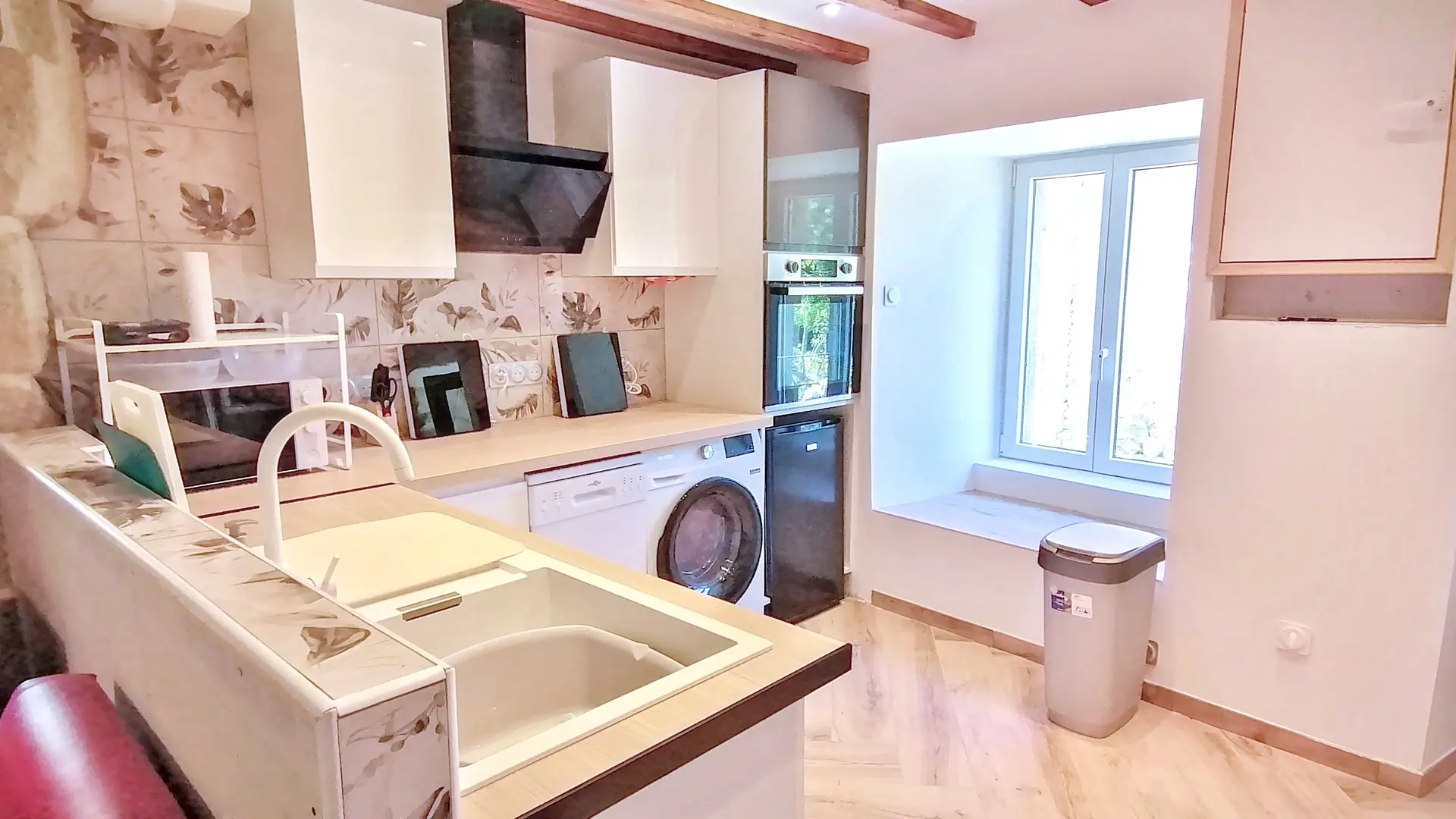 Une vue en gros plan d'une cuisine moderne de style campagnard, contrastant avec des éléments rustiques. Au premier plan à gauche, un plan de travail blanc et brun foncé avec un grand évier encastré est visible. Une partie du mur arrière est carrelée de carreaux à motifs floraux beiges et marron. À droite, une fenêtre laisse entrer la lumière naturelle et dispose d'un large rebord. Le plafond est doté de poutres en bois apparentes. À l'extrême gauche, on aperçoit une partie d'un mur en pierres rustiques.