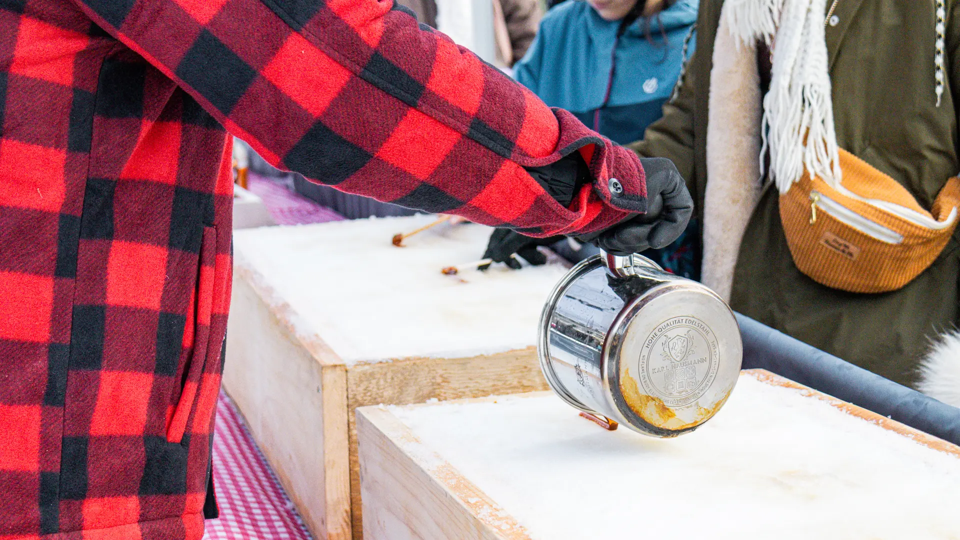 Une personne portant une veste à carreaux rouges et noirs verse du sirop chaud sur de la neige fraîchement tassée dans des bacs en bois pour fabriquer de la tire sur neige.