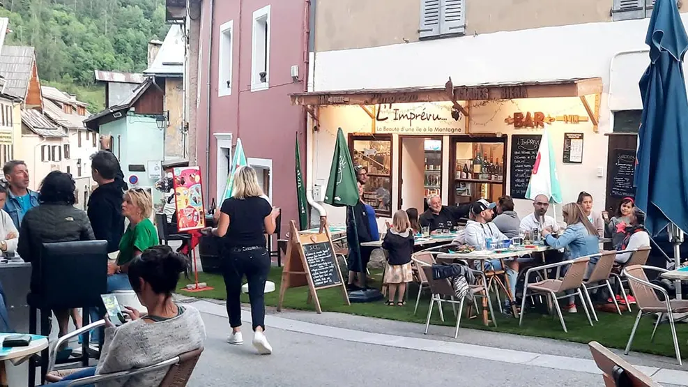 Terrasse extérieure du restaurant, située dans la grand rue d'Allos, fausse pelouse au sol, tables et chaises. Inscription sur façade : 'L'Imprévu, La Beauté d'une île à la montagne