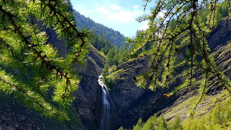 Cascade visible en fond au milieu d'une formation rocheuse entourée par la végétation. Montagnes boisées en arrière-plan