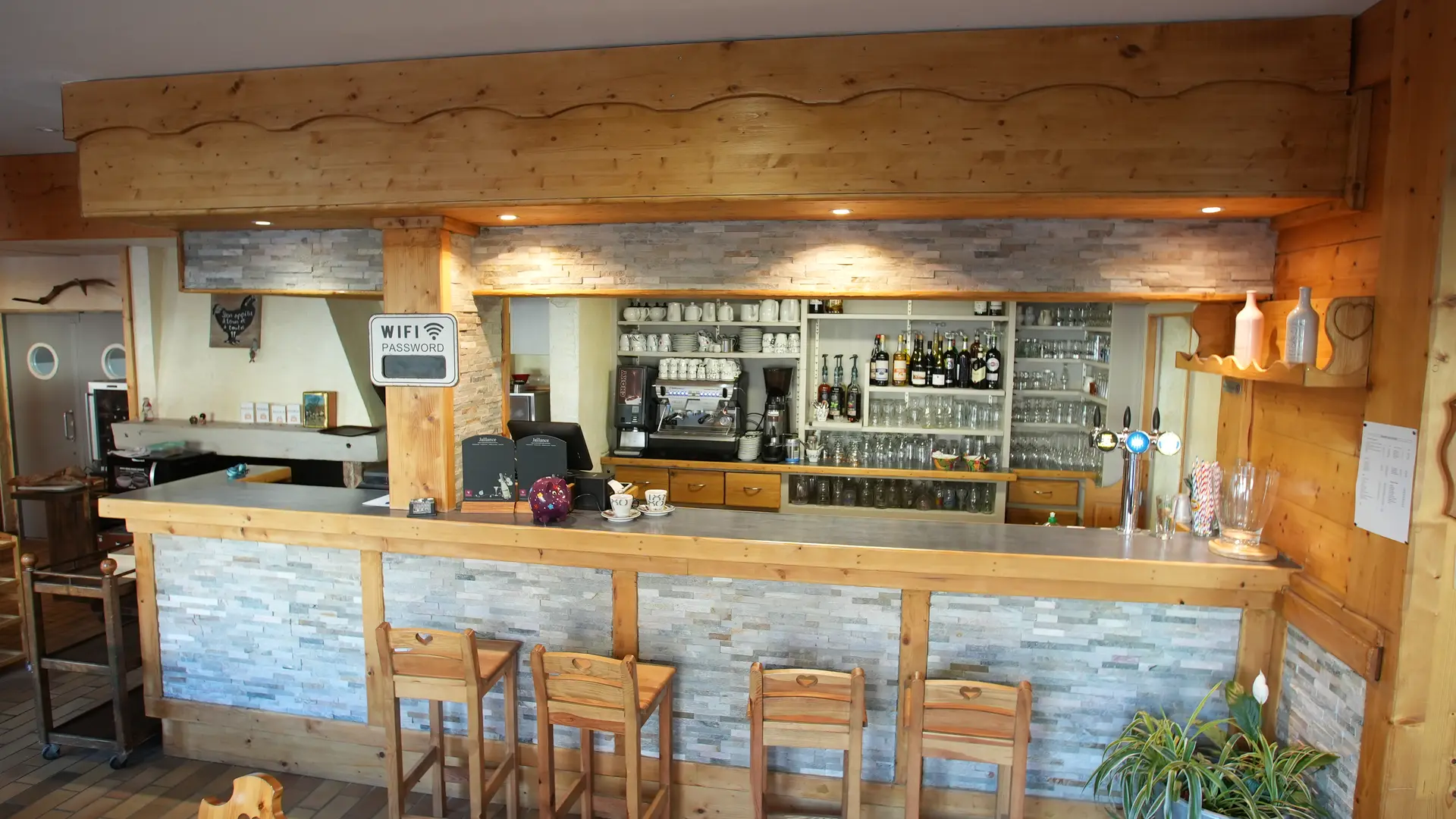 Le bar dispose de quatre tabourets. Sur les étagères murales sont rangés bouteilles, verres, tasses et machine à café/thé. Une plante verte occupe l’angle droit et le code Wi-Fi est affiché