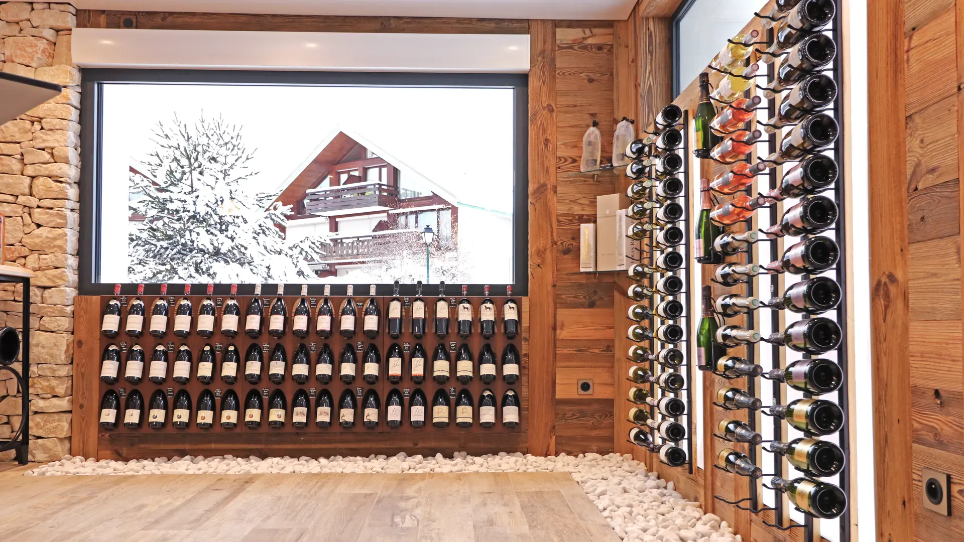 Étagères cave à vins avec de nombreuses bouteilles et en fond la vue sur un chalet enneigé depuis la fenêtre.