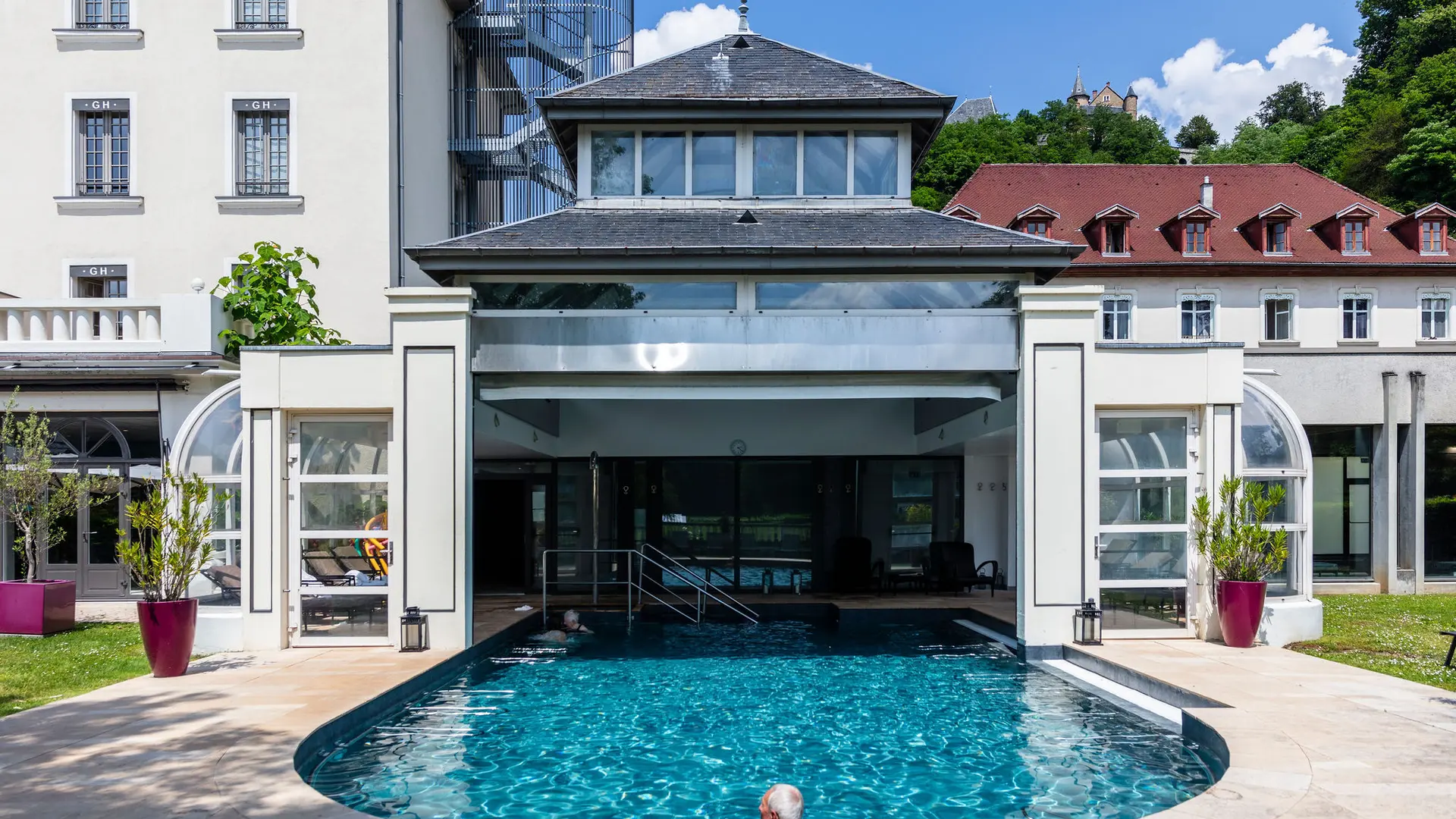 L'image présente un espace extérieur avec une piscine dans un cadre verdoyant. À l'arrière-plan, on aperçoit le grand hôtel et thermes d’Uriage, des lignes droites et des fenêtres régulières.