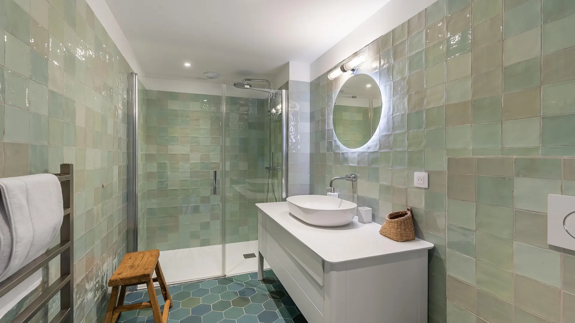 Salle de bains avec douche à l'tialtienne, vasque et meuble, miroir rond, briques de verre aux murs, sèche serviettes