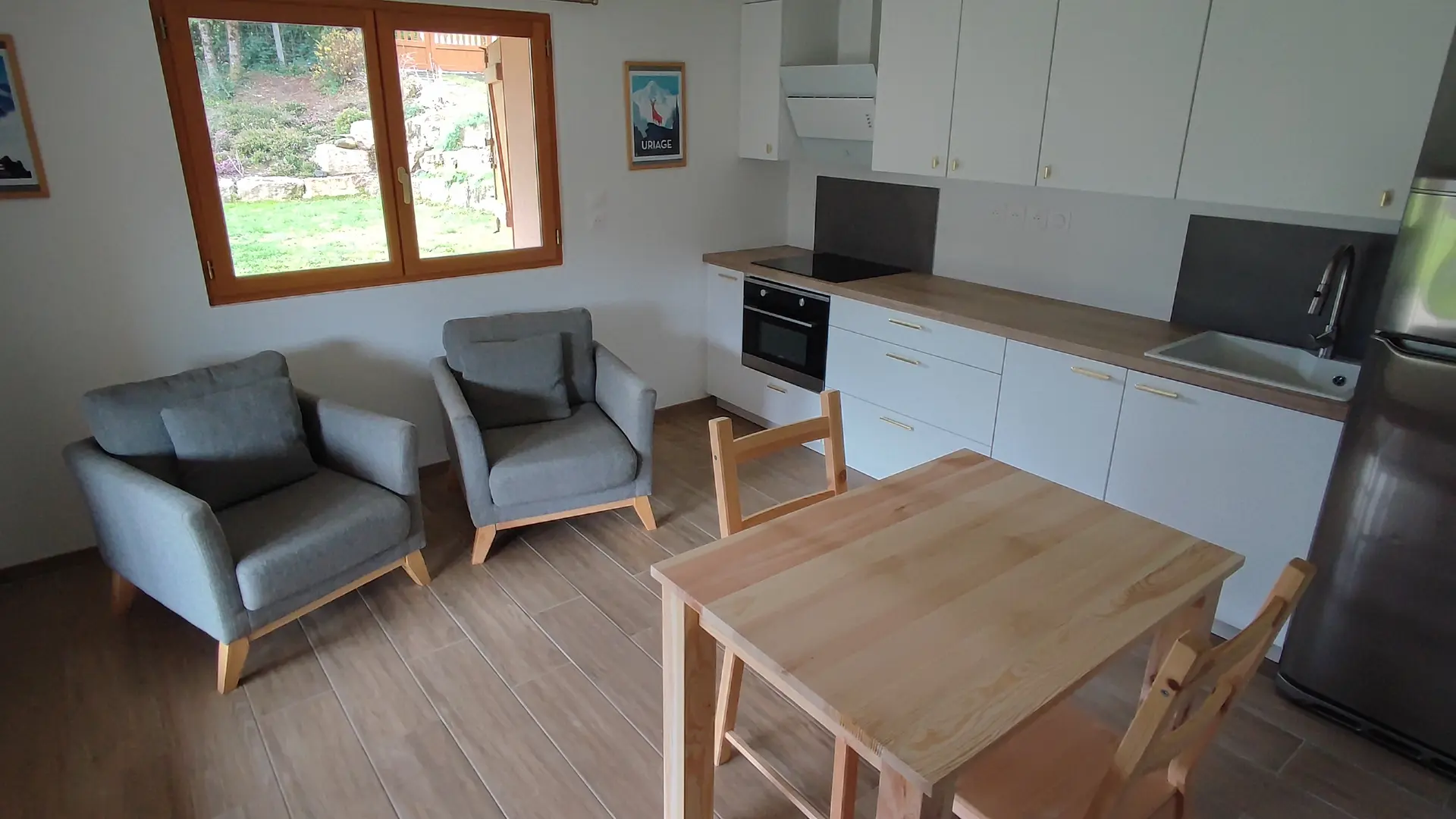 Cette pièce de vie réunit une kitchenette avec un long plan de travail blanc, un coin repas central avec une table en bois et deux fauteuils gris disposés sous une fenêtre.