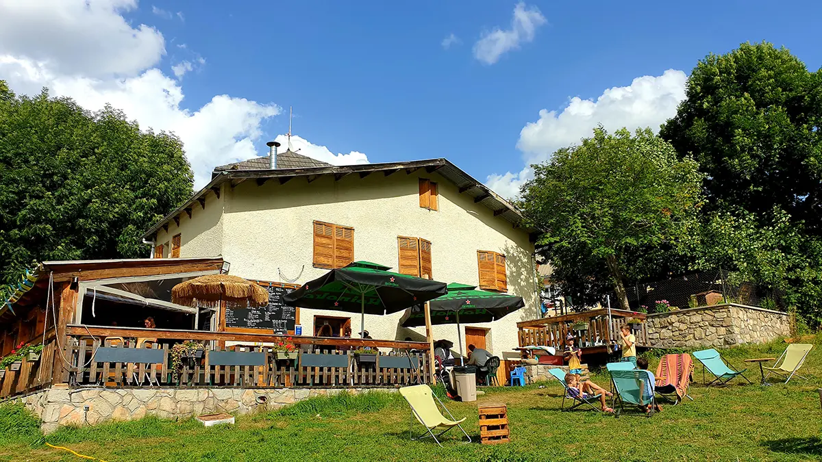 Overview of the restaurant, a two-story stone and wood building with a wooden terrace on the ground floor, tables, chairs, parasols, and sun loungers. Meadow in front of the restaurant with sun loungers.