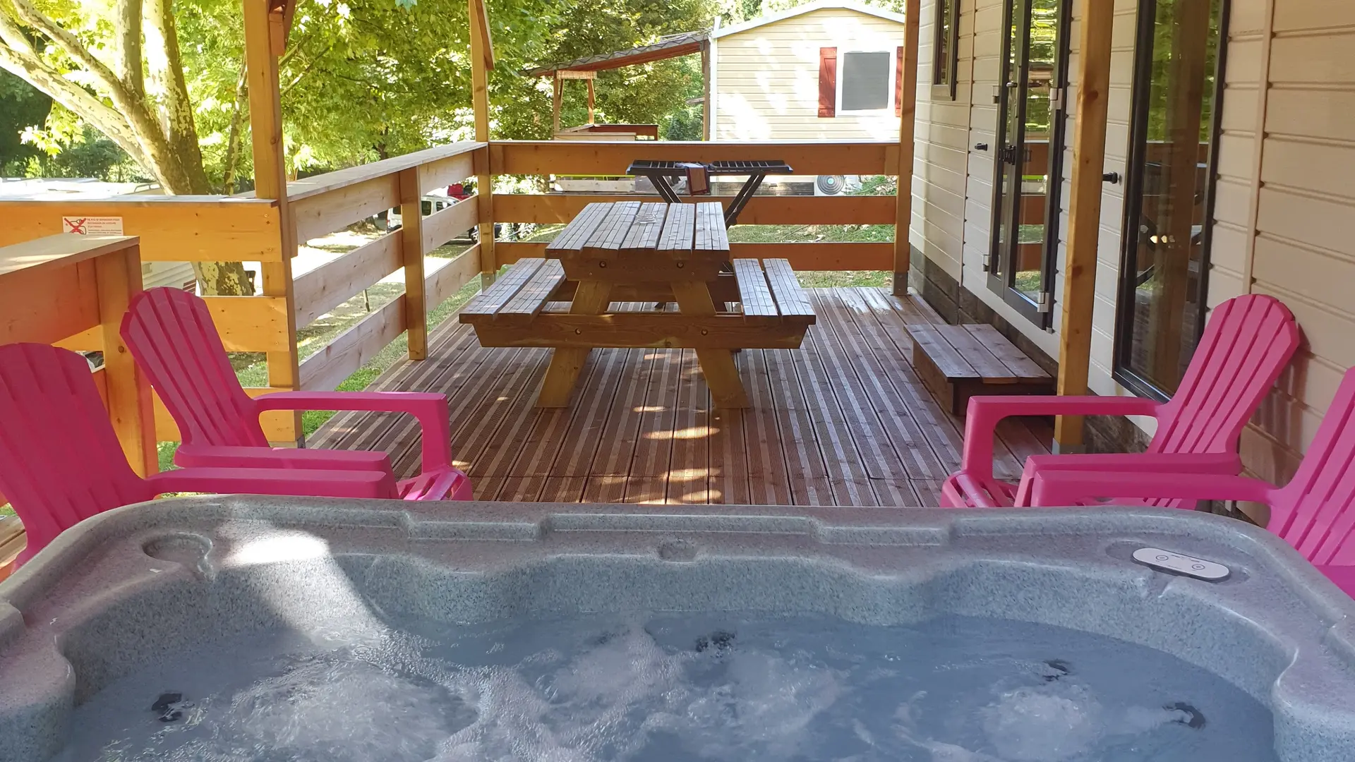 L’extérieur du mobil-home aménagé avec un jacuzzi, une terrasse en bois couverte, des chaises longues et une table de pique-nique.