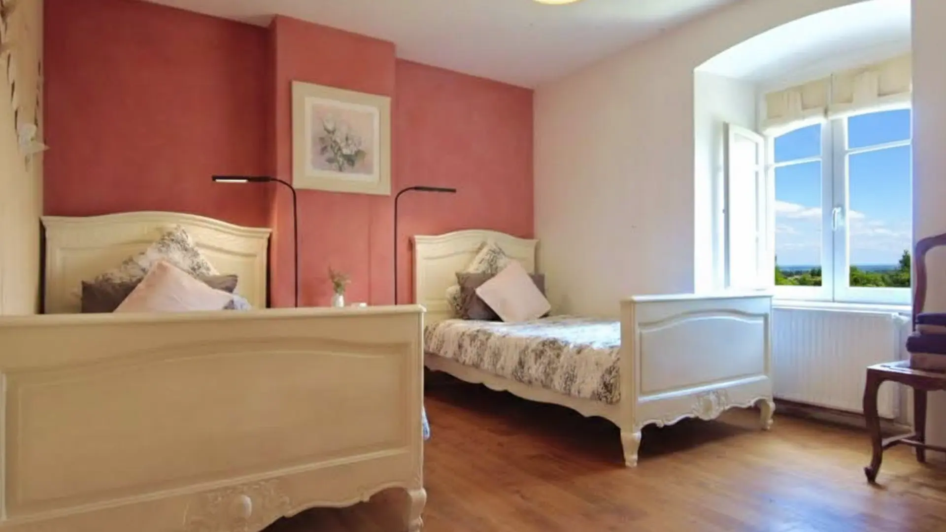Une chambre romantique avec deux lits simples anciens et revisités vous accueillent dans cette chambre lumineuse. Le mur à droite est de couleur terracota avec un tableau séparant les lits. Les lits sont blancs et à la tête de lit 3 coussins assortis au jeté de lit vous attendent pour un repos bien mérité. Une grande fenêtre s'ouvre sur un ciel bleu azur et une végétation abondante. Il y a une chaise ancienne à droite de la fenêtre. Le sol est un beau plancher chaleureux.