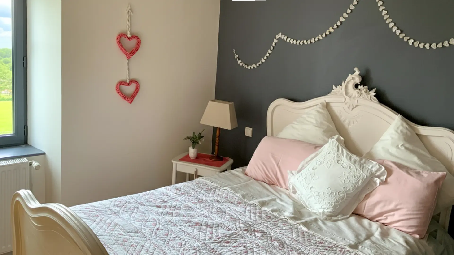 C'est la troisième chambre, toujours dans un style romantique avec son ancien lit. Le mur de la tête de lit est gris foncé et il contraste avec la banderole de coeurs  au-dessus. Le reste des murs sont blancs. A côté du lit, il y a une table de chevet avec sa lampe et une petite plante. Sur le mur, sont accrochés deux coeurs rouges contrastant avec le mur blanc. A la gauche du mur, une fenêtre apporte une belle luminosité à la chambre.
