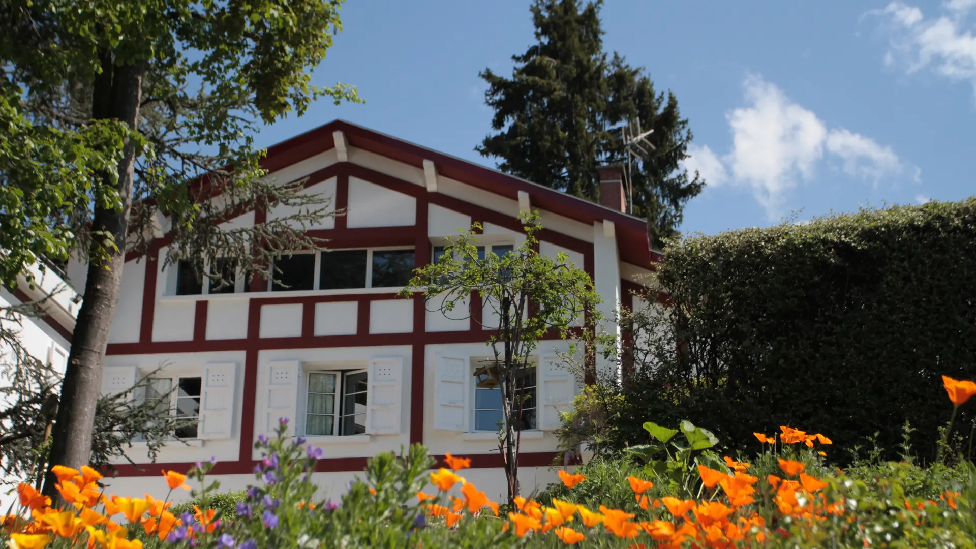 Cette maison de style traditionnel possède un toit à deux pans et des murs à colombage. Elle est entourée d'un jardin où l'on peut voir de nombreuses fleurs colorées.