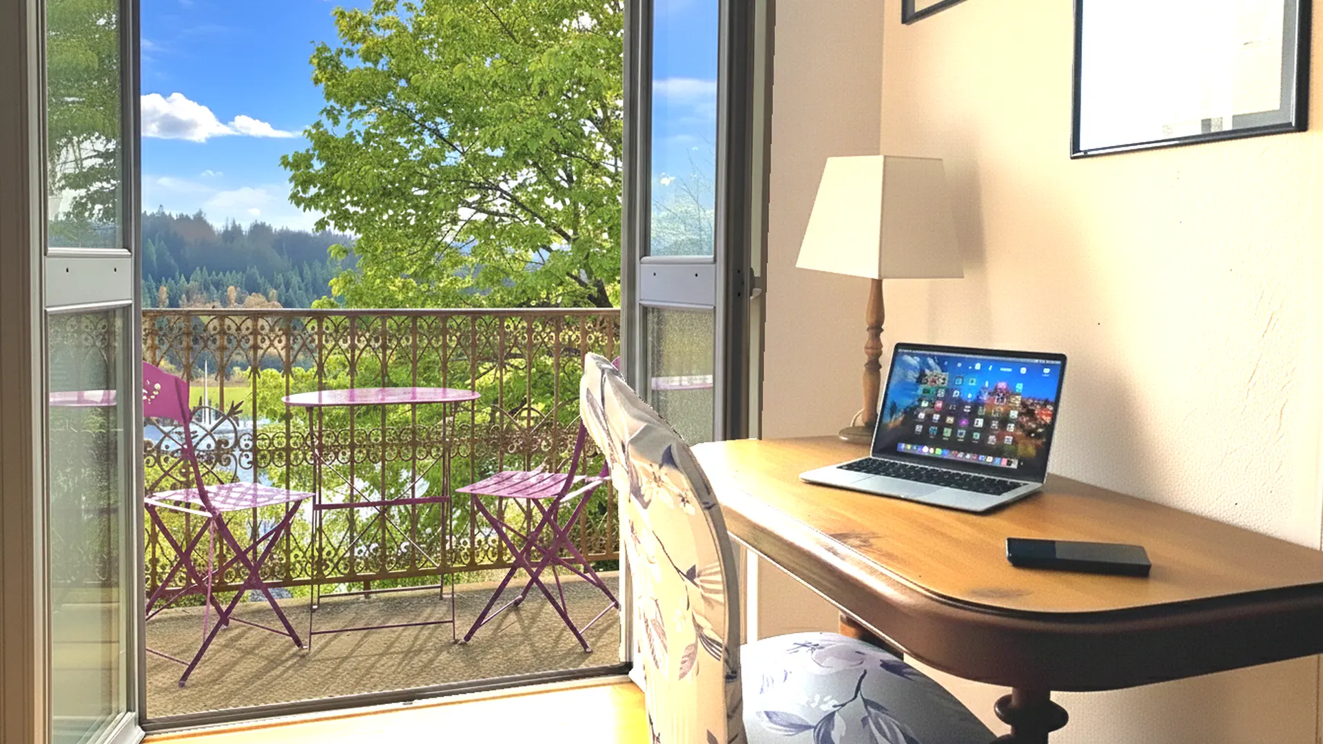 Petit espace bureau lumineux donnant sur un balcon. Un ordinateur portable est posé sur un bureau en bois, accompagné d’une lampe à pied et d’un téléphone. Le balcon, accessible par une grande porte-fenêtre ouverte, est aménagé avec une petite table ronde et deux chaises pliantes violettes. La balustrade en fer forgé s’ouvre sur une vue dégagée : arbres verts, collines boisées et ciel bleu parsemé de nuages. L’ensemble crée une atmosphère paisible et propice au travail comme à la détente.