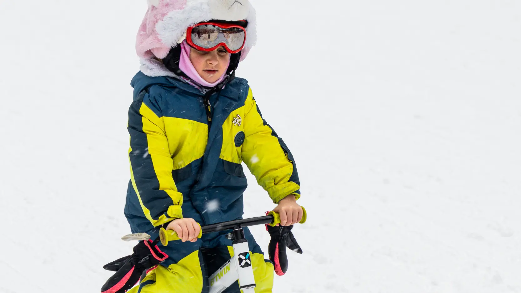 Voici une description simple pour cette dernière image, suivie de ses traductions : Un jeune enfant portant une combinaison de ski colorée et un bonnet rose en forme de tête d'animal descend une pente de neige sur un petit vélo-ski blanc.