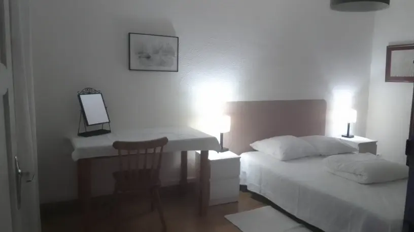 une chambre à coucher avec un lit au centre, une tête de lit en bois clair, des tables de chevet avec des lampes allumées, et un bureau ou une coiffeuse sur la gauche.
