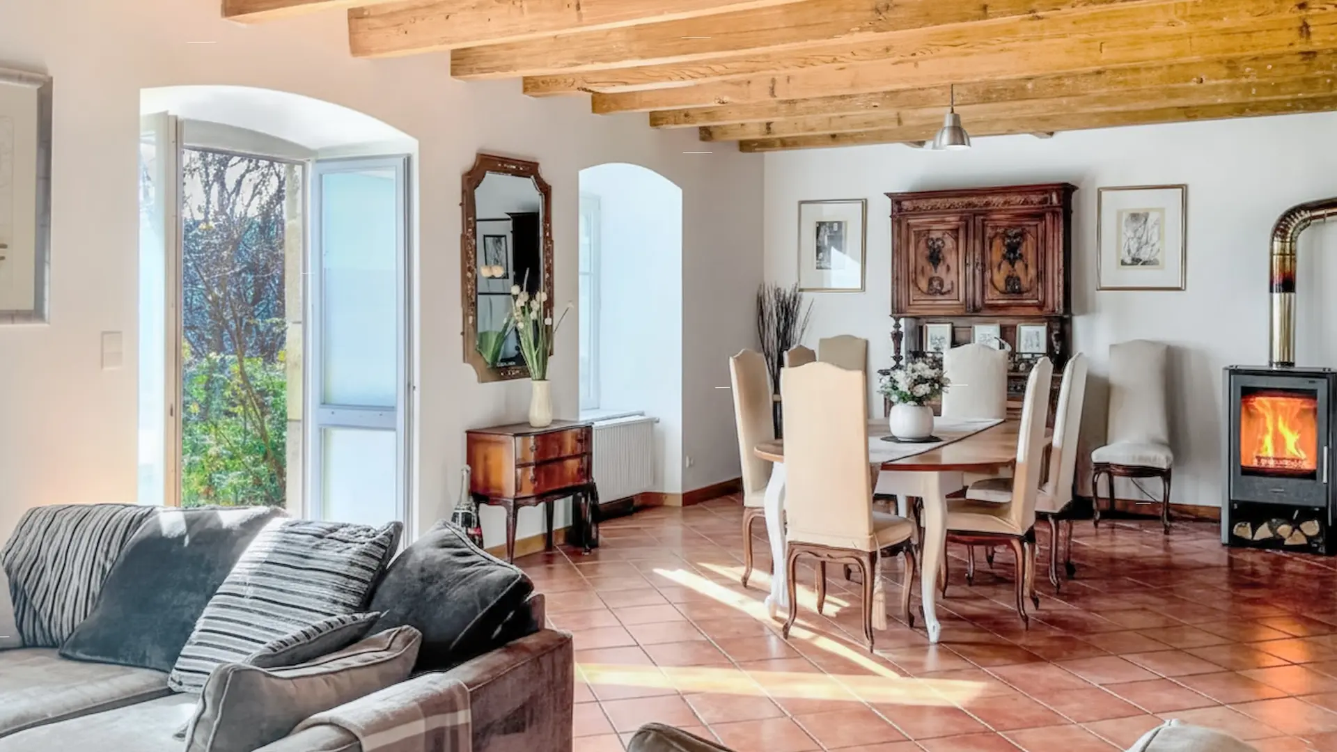 Grand séjour lumineux mêlant confort moderne et charme traditionnel. Au premier plan, un large canapé gris avec des coussins rayés et un plaid invite à la détente. Plus loin, une salle à manger élégante occupe l’espace : table ronde en bois clair entourée de chaises hautes tapissées, et un buffet ancien en bois sculpté contre le mur. Un poêle à bois diffuse une lumière chaude, soulignant le sol en carreaux terracotta. Le plafond est traversé de poutres en bois naturel. De grandes portes-fenêtres ouvertes laissent entrer la lumière du jour et révèlent la verdure extérieure. L’ensemble respire la douceur, la chaleur et la convivialité.
