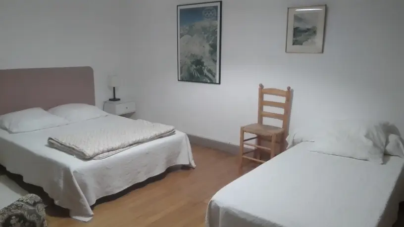 Cette chambre présente deux lits un simple et un double avec du linge de lit blanc, un fauteuil en bois entre les deux lits, et des tableaux sur le mur.
