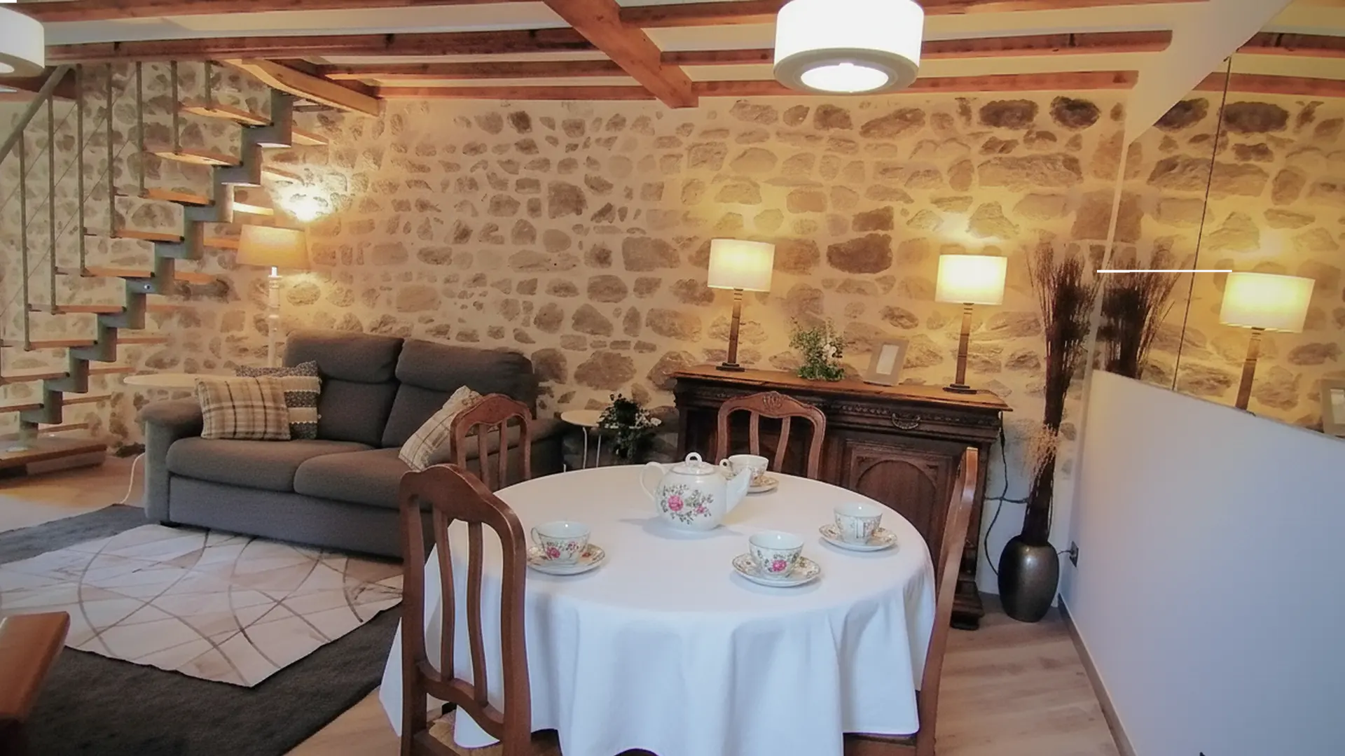 Un coin repas et salon chaleureux dans un espace ouvert. Le mur du fond est recouvert de pierres apparentes, créant une ambiance rustique. Le plafond est orné de poutres en bois.  Au premier plan, une petite table ronde est recouverte d'une nappe blanche et dressée avec une théière et des tasses à motifs floraux. Elle est entourée de trois chaises en bois de style classique.  Contre le mur de pierres, entre le coin repas et le salon, se trouve un petit buffet en bois sombre de style antique, surmonté de deux lampes de table avec des abat-jours carrés beiges. Un grand miroir mural à droite reflète cette scène.  Dans le coin salon, à l'arrière-plan, un canapé gris est agrémenté de coussins à carreaux. Un tapis gris à motifs géométriques clairs délimite cet espace. Un escalier ouvert avec des marches en bois et un limon métallique monte le long du mur gauche, menant à un autre niveau.