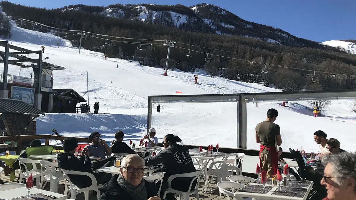 Terrasse extérieure du restaurant en hiver, tables et chaises en plastique, vue sur les pistes enneigées et les montagnes, télésiège à proximité