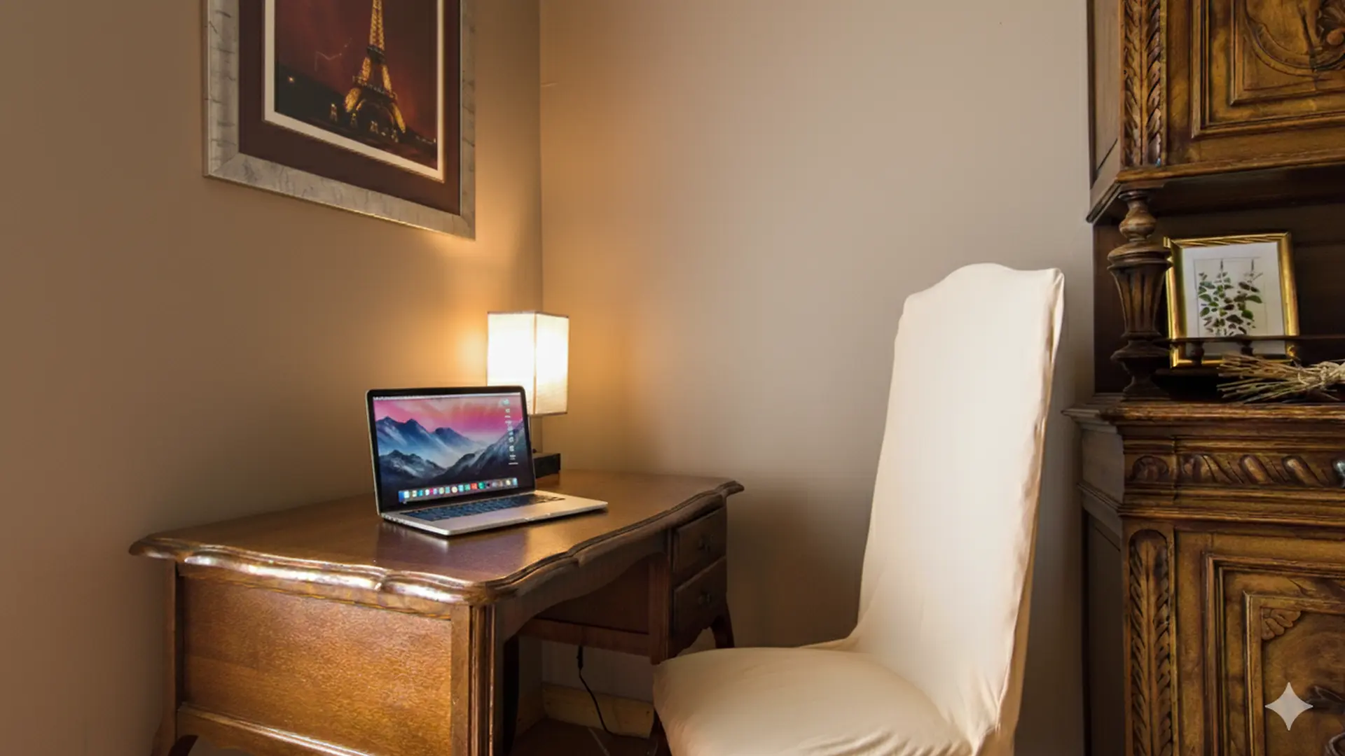 Petit espace bureau chaleureux avec un bureau en bois ancien sur lequel repose un ordinateur portable ouvert. Une lampe douce éclaire le coin de travail. Une chaise recouverte d’un tissu clair est placée devant le bureau. À droite, un meuble en bois sculpté ajoute une touche traditionnelle, décoré d’un petit cadre et de quelques branches. Une photo encadrée de la tour Eiffel est accrochée au mur, donnant une ambiance élégante et intime.