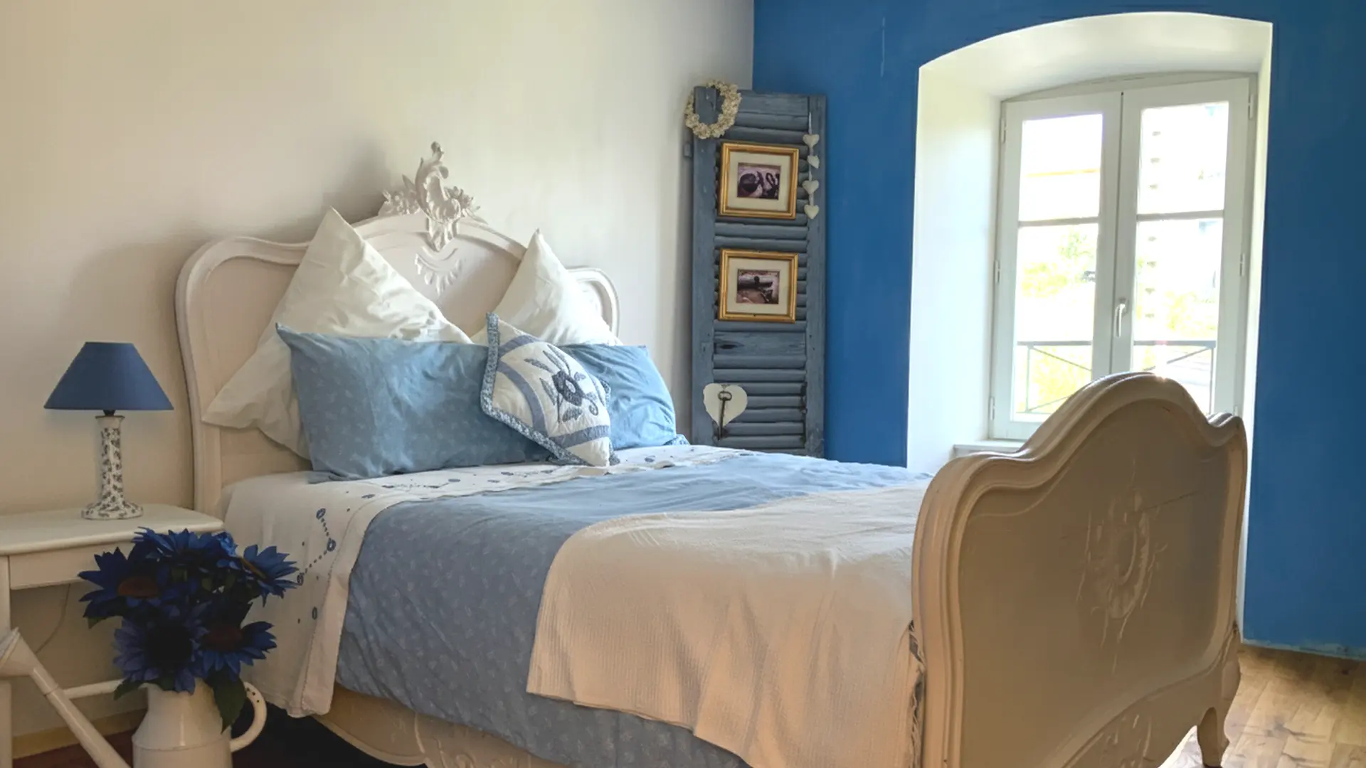 Chambre lumineuse avec un lit ancien en bois sculpté de couleur crème, recouvert d’une literie bleue et blanche et de plusieurs coussins assortis. À côté du lit, une petite table blanche porte une lampe à abat-jour bleu et un vase rempli de fleurs bleues. Le mur derrière la tête de lit est clair, tandis qu’un mur bleu vif entoure une grande fenêtre à deux battants laissant entrer beaucoup de lumière naturelle. Une décoration composée de cadres et de cœurs en bois est accrochée sur un vieux volet bleu à droite du lit, donnant une ambiance douce et bord de mer.