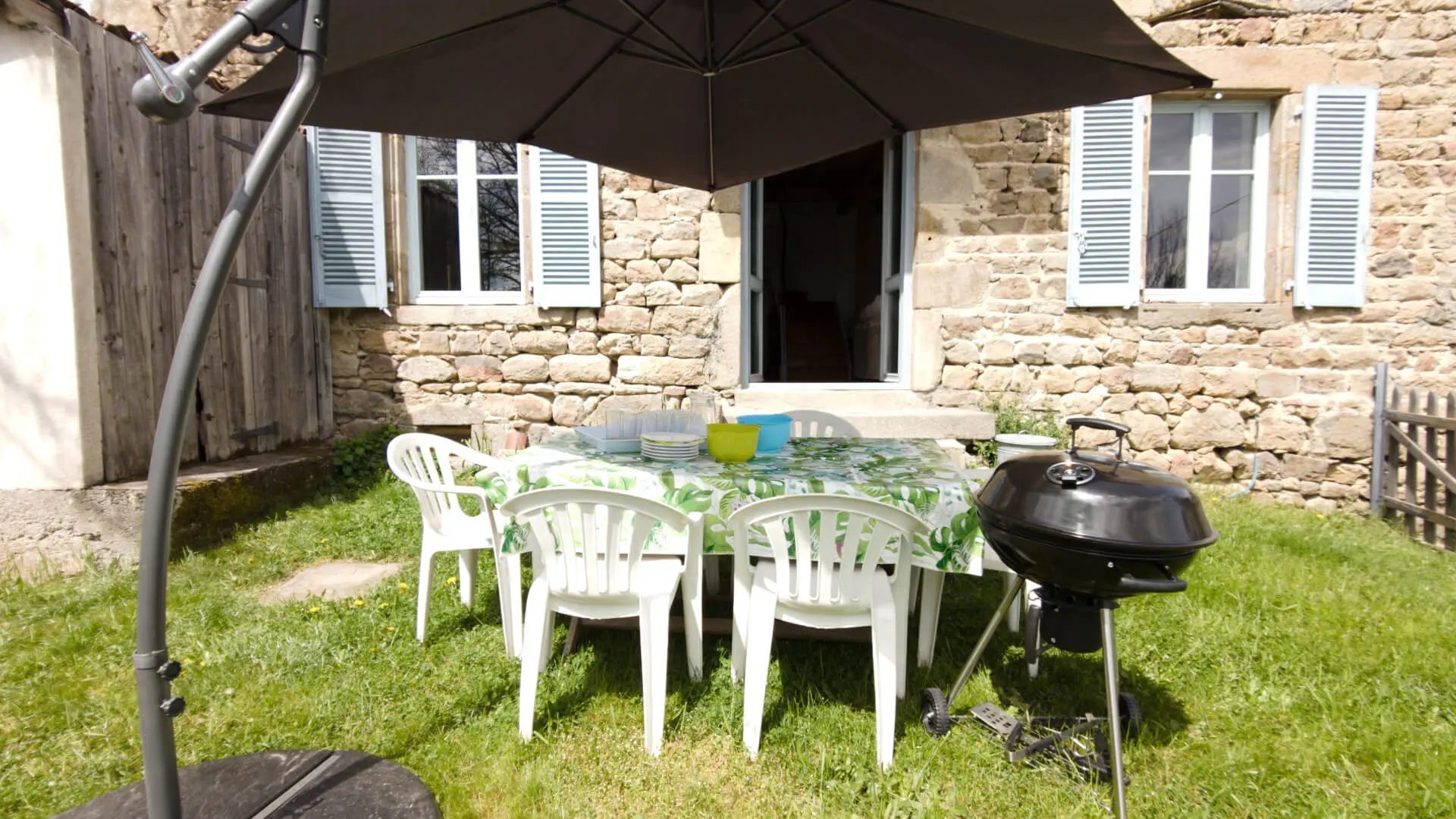 Un accès à l'extérieur est possible par deux -trois marches en pierre. La porte est positionnée au salon. Le jardin est herbeux avec sur la gauche en sortant du gîte, un portillon pour fermé le jardin; On trouve une table et des chaises de jardin blancs. La table a une nappe avec des feuillages sur fond blanc qui s'intègre dans le paysage.  Un parasol décentré de couleur gris protège du soleil la table; un barbecue grill rond est à votre disposition.