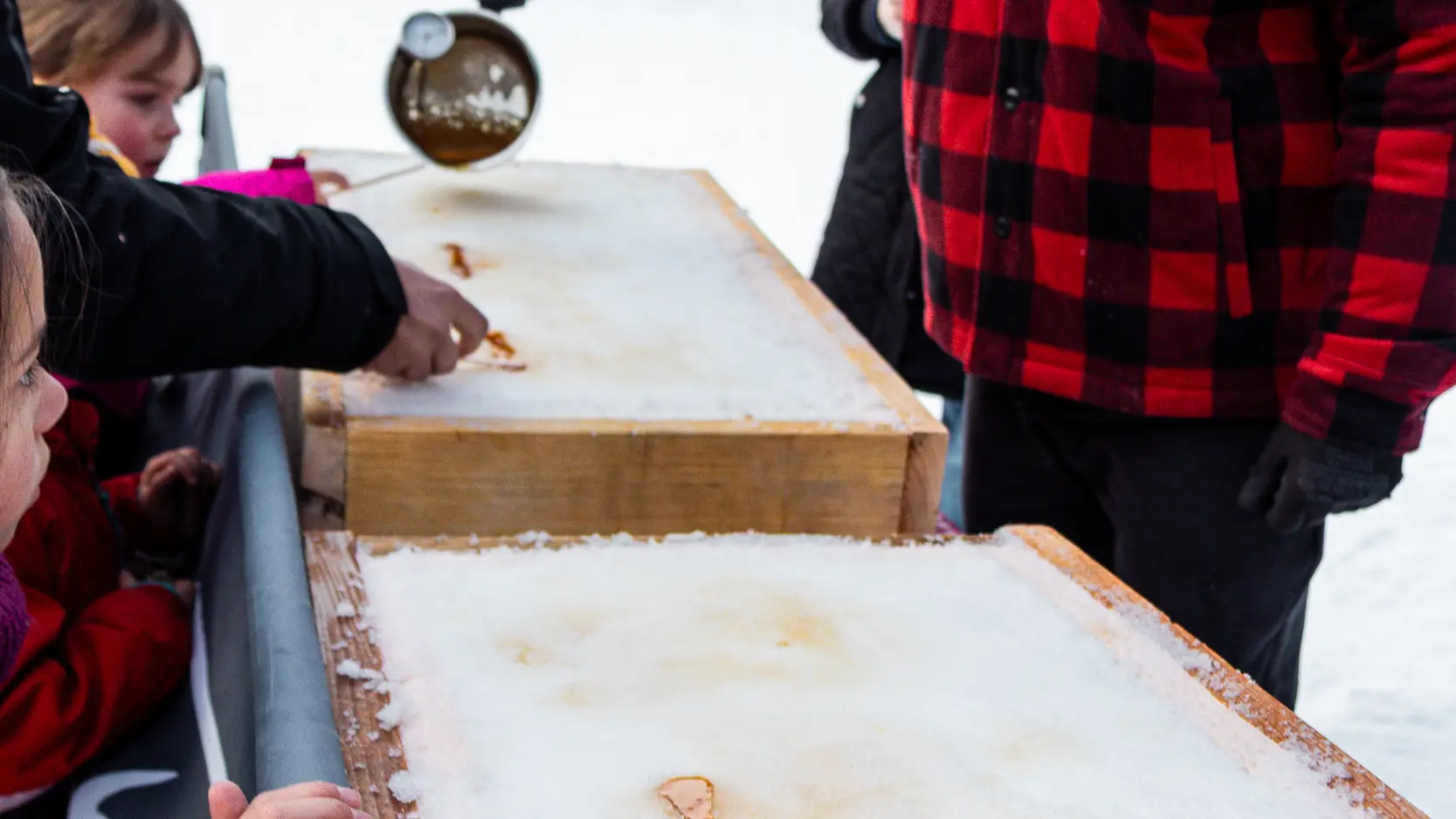 On voit des enfants s'initier au vélo-ski sur la neige avec l'aide d'adultes. À côté, une animation gourmande permet de déguster de la tire sur neige en enroulant du sirop chaud sur des bâtonnets.