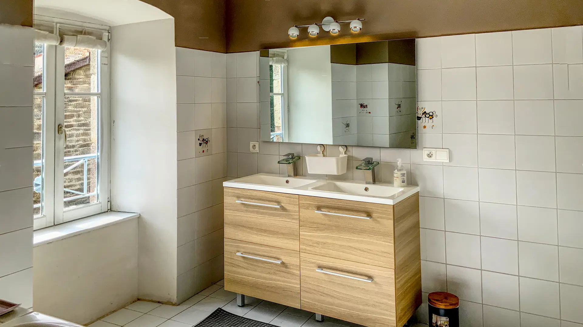 Une salle de bain lumineuse avec des murs et un sol carrelés de blanc. Au centre, un meuble-lavabo moderne en bois clair avec quatre tiroirs et deux éviers intégrés. Au-dessus du meuble-lavabo, un grand miroir rectangulaire est éclairé par une rampe de quatre ampoules rondes. Des carreaux décoratifs avec de petits motifs colorés sont visibles sur le mur derrière les éviers. À gauche, une fenêtre à petits carreaux, encadrée de blanc, est ouverte et donne sur l'extérieur. Un rebord de fenêtre en plâtre blanc est visible en dessous. Le plafond est marron et une partie d'une baignoire est visible dans le coin inférieur gauche de l'image. Un petit tabouret marron et une poubelle noire sont dans le coin inférieur droit.
