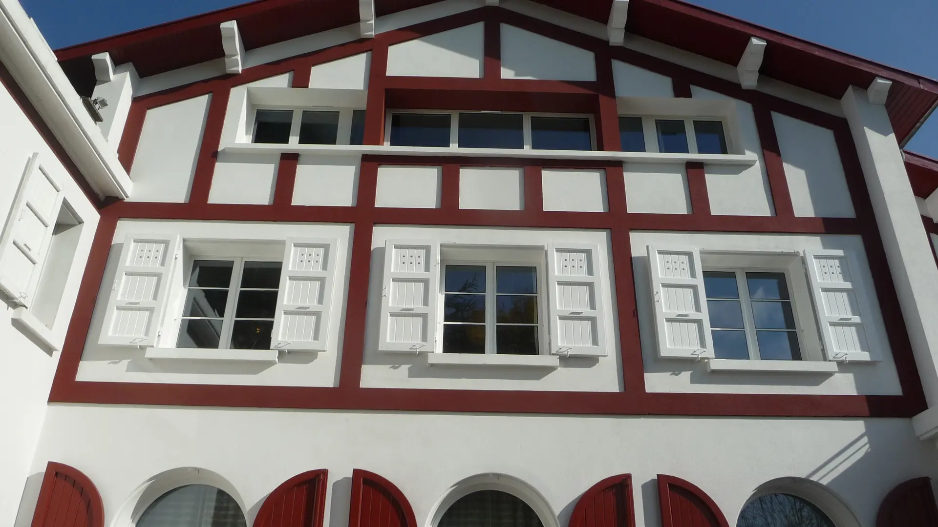 Cette maison possède deux étages et un rez-de-chaussée. Ses murs sont blancs et ses poutres en bois apparentes sont de couleur rouge. De grandes fenêtres dotées de volets rouges donnent sur l'extérieur.