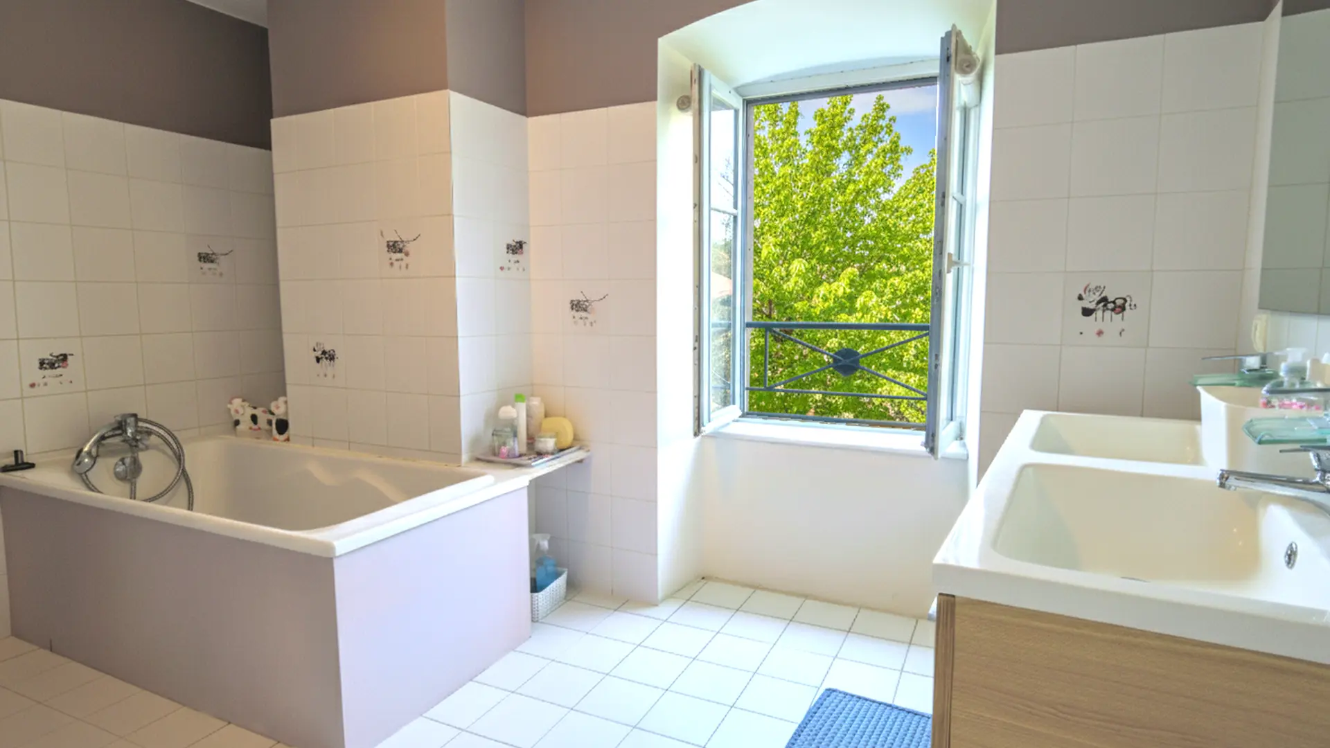 Une salle de bain lumineuse avec des murs carrelés de blanc et un sol carrelé de blanc. Une baignoire rectangulaire est située à gauche, avec un tablier de couleur lavande pâle. Le carrelage blanc des murs est décoré de petits motifs fantaisistes. Un robinet de douche chromé est fixé au mur au-dessus de la baignoire. Près de la baignoire, une petite étagère en bois clair contient des produits de bain. À droite, on aperçoit une partie d'un meuble-lavabo en bois clair avec deux éviers blancs. Une fenêtre ouverte au centre de l'image laisse entrer la lumière naturelle et offre une vue sur une verdure luxuriante. La fenêtre est sécurisée par une balustrade en fer forgé. Le plafond est de couleur gris-brun. Un petit tapis de bain bleu est posé sur le sol devant les éviers.