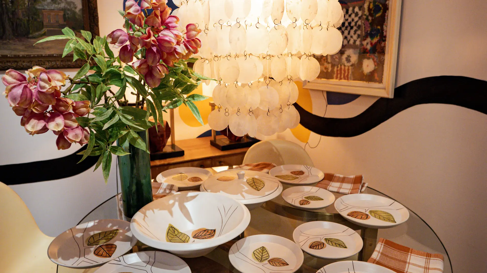 Une table ronde élégamment dressée mêle vaisselle vintage et ambiance chaleureuse sous un lustre en nacre.