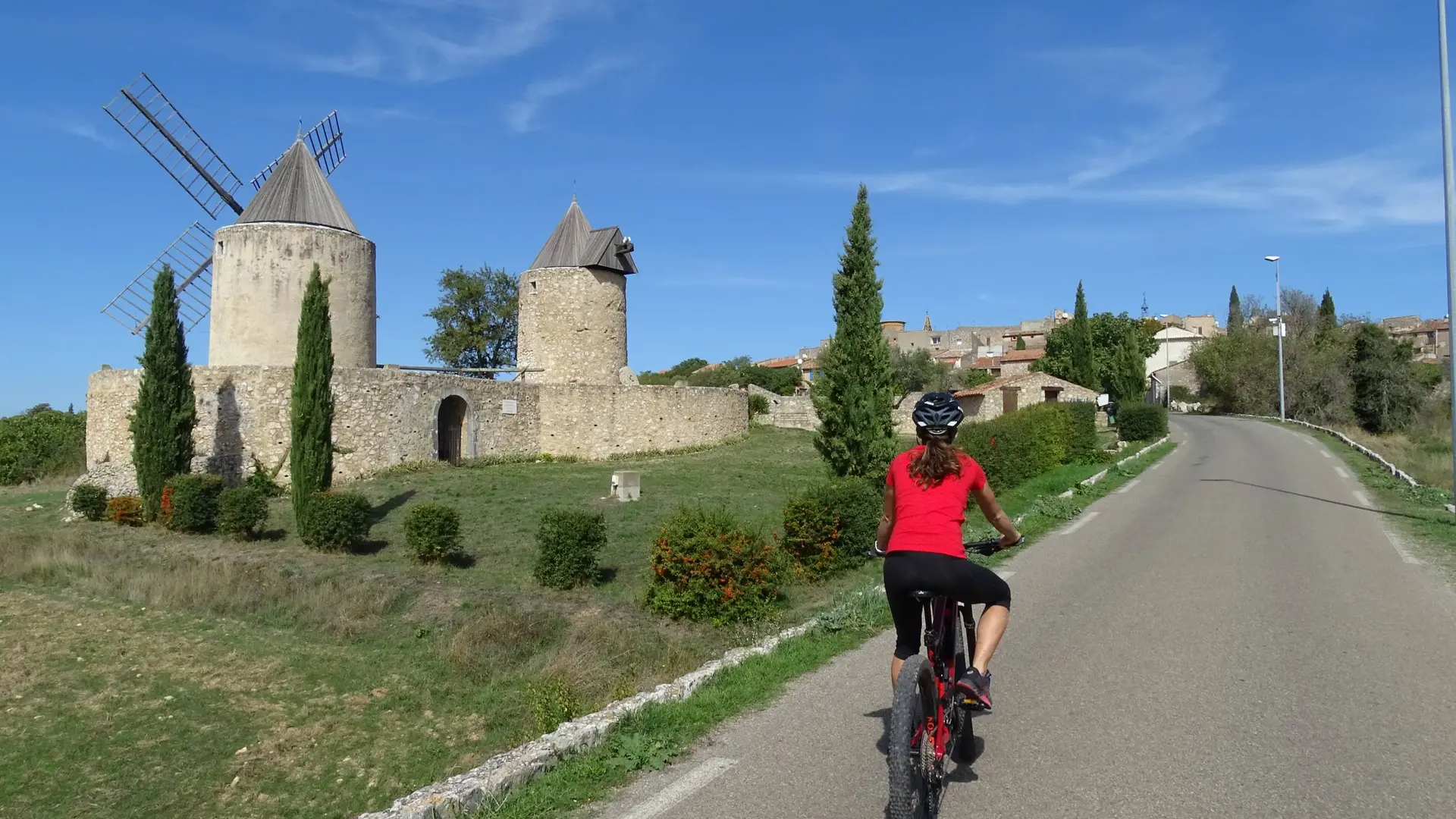 Cycliste sur route goudronnée avec en deuxième plan les 2 moulins en pierres à l'entrée d'un pré