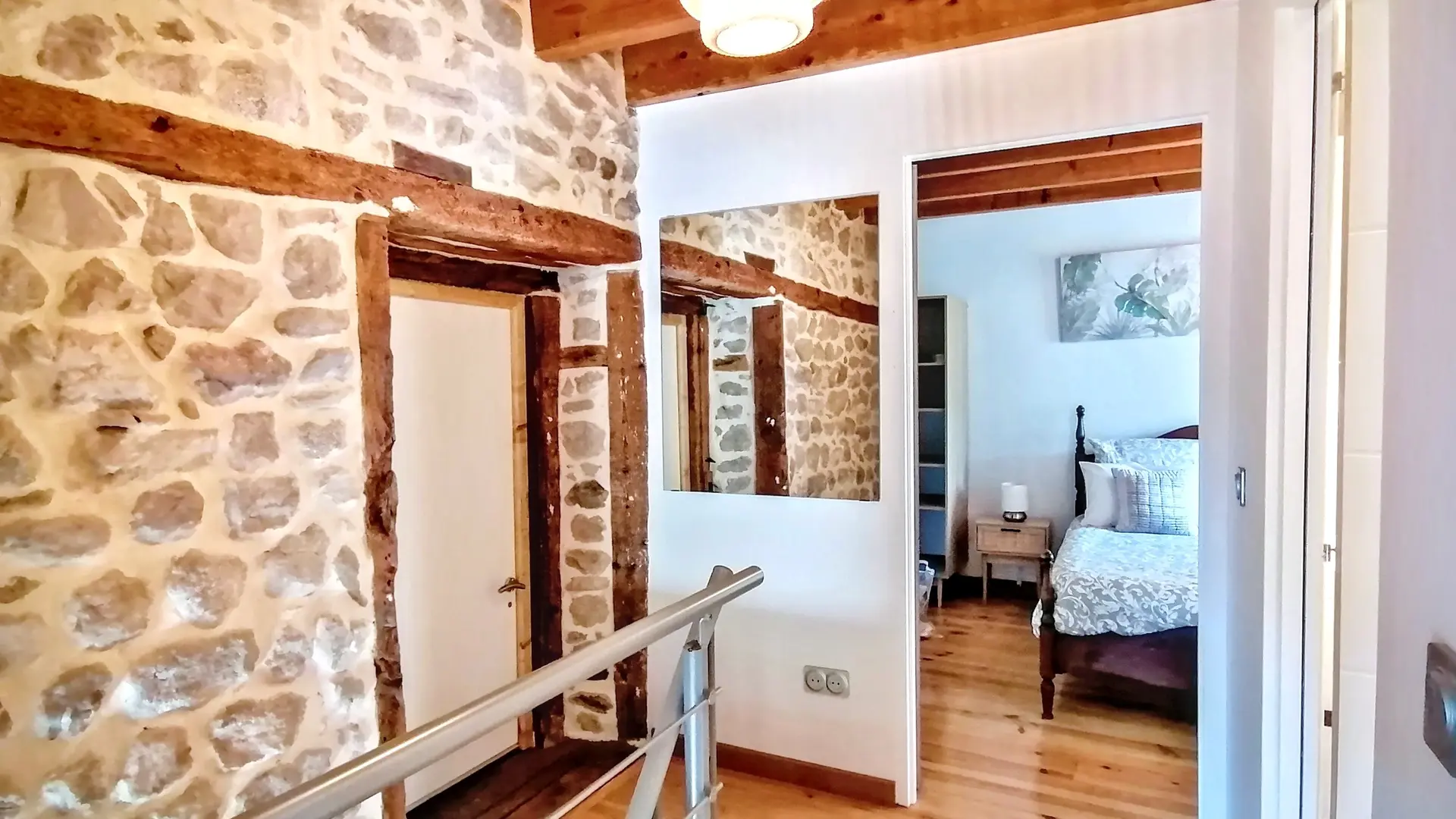 Un palier à l'étage dans une maison mêlant style rustique et moderne. À gauche, un mur est constitué de pierres apparentes, traversé par des poutres en bois horizontales et verticales qui encadrent une porte blanche fermée. Une rampe d'escalier métallique et chromée avec des montants fins est visible au premier plan. Le sol est un plancher de bois clair, et le plafond est orné de poutres en bois.  Au centre et à droite, un mur blanc présente un miroir rectangulaire qui reflète les poutres et les pierres. Une ouverture dans le mur mène à une chambre à coucher. À travers cette ouverture, on aperçoit un lit double à structure en bois sombre et une literie blanche. Un tableau floral est accroché au mur au-dessus du lit, et une table de chevet en bois clair est visible à côté du lit.