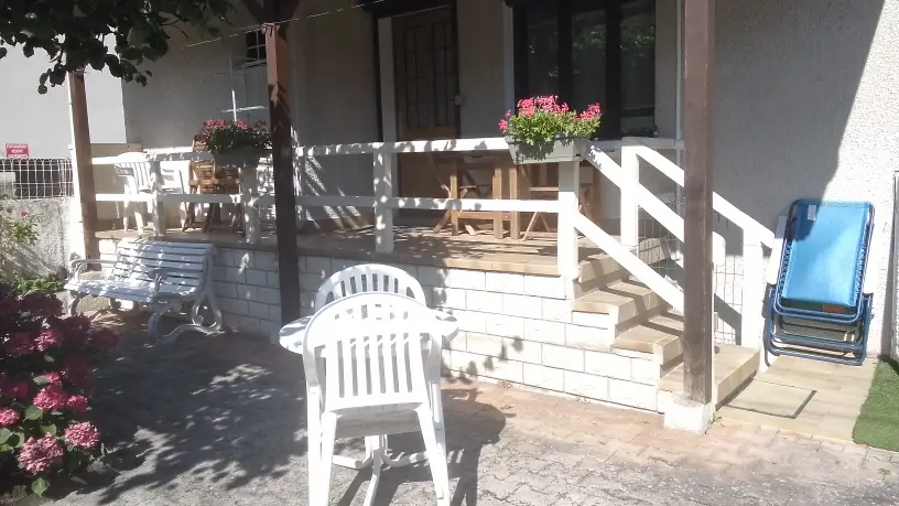 Une chaise blanche en plastique au premier plan sur une terrasse, faisant face à un escalier et une balustrade en bois qui mènent à une véranda sombre ou une entrée de maison.
