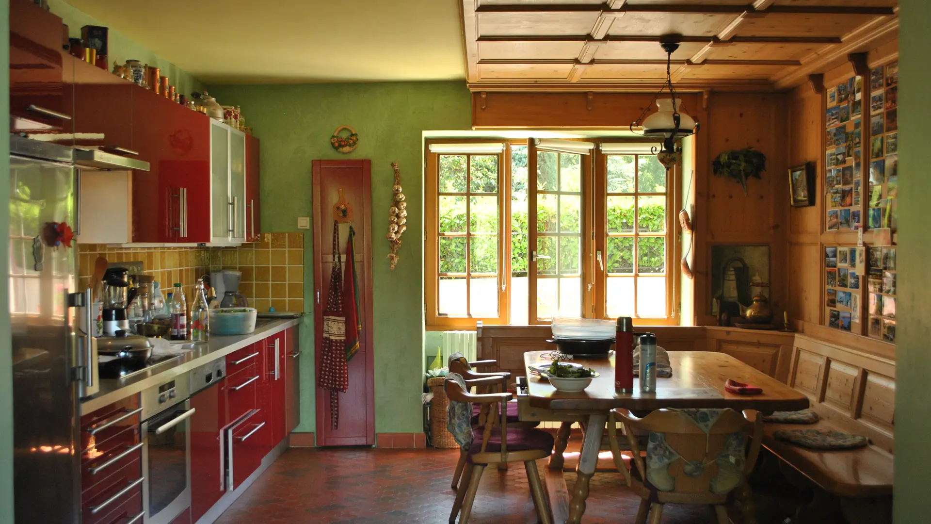 Vue sur une cuisine assez spacieuse et chaleureuse dans les tons rouge, vert et bois. Présence d'une grande cuisine aménagée à gauche de la photo et un espace pour manger tout en bois sur la droite. Au fond de la pièce, se trouve une large fenêtre qui donne sur le jardin